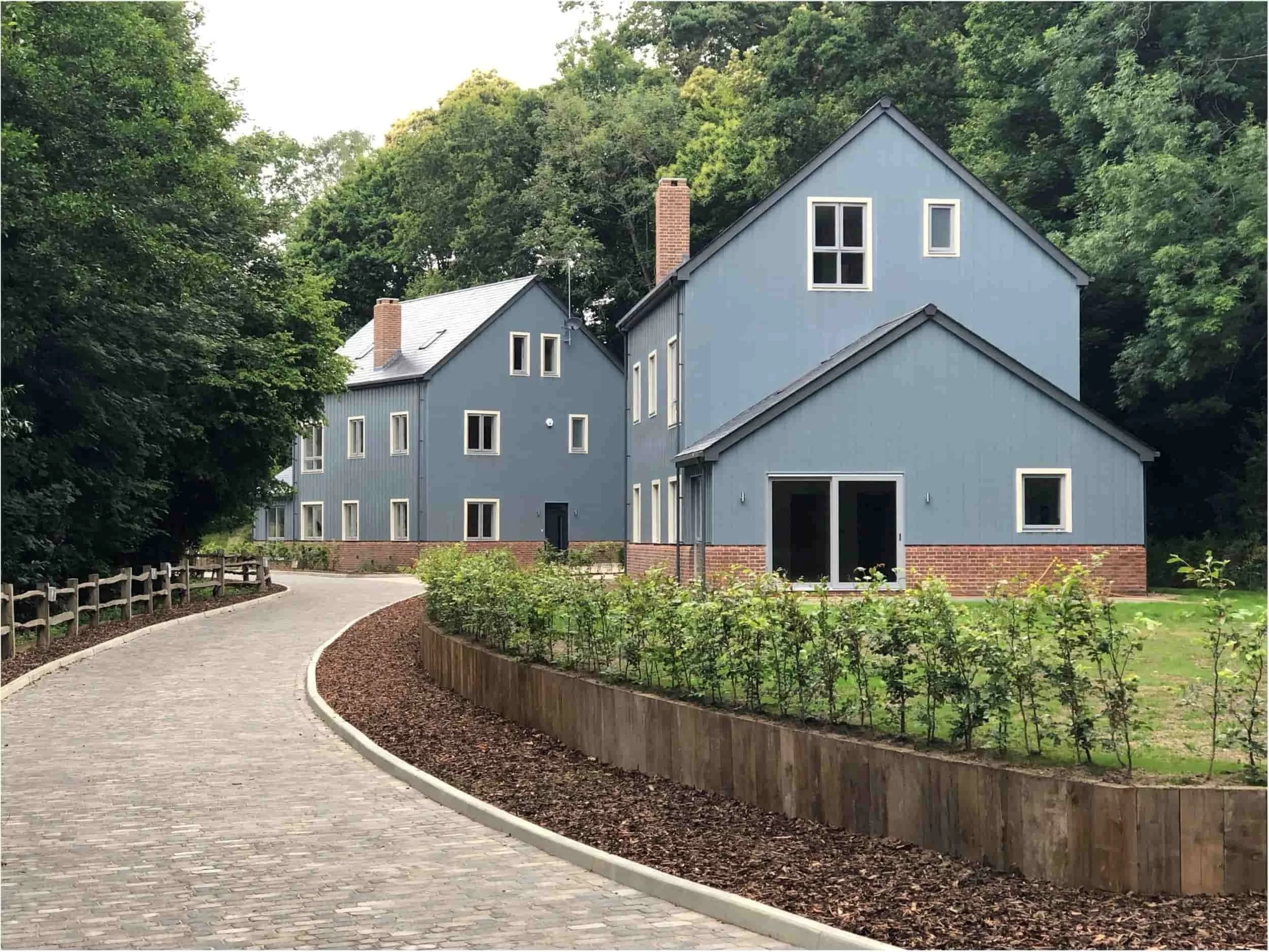 Modern blue-gray multi-building house with brick accents, landscaped garden, and curved cobblestone driveway surrounded by trees