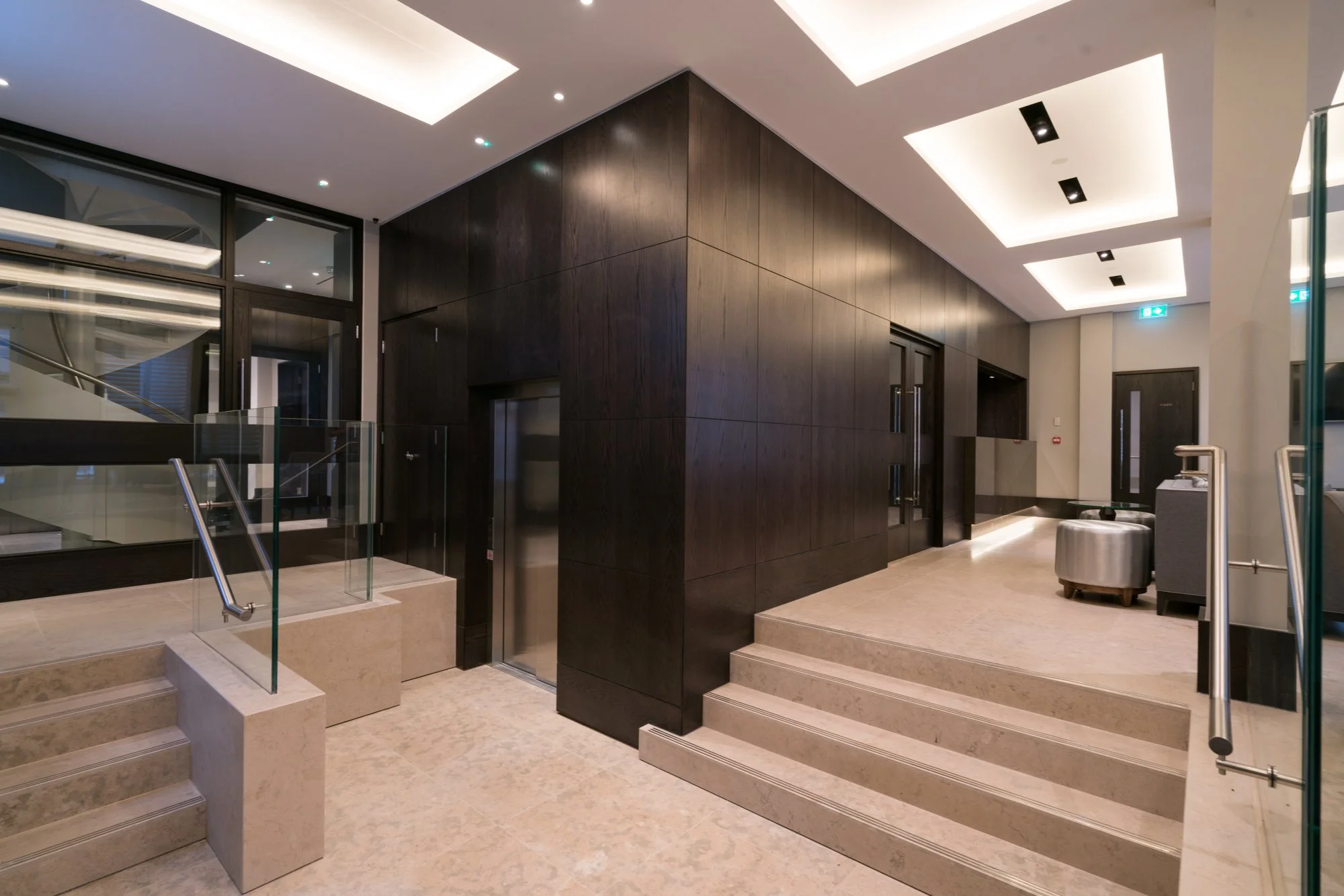Modern luxury building lobby with dark wood-paneled walls, marble steps, glass railings, elevator, and recessed ceiling lighting