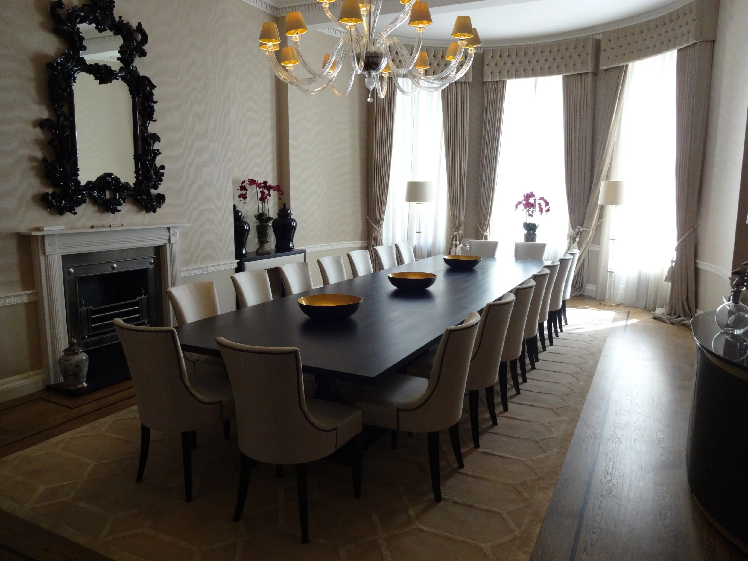 Elegant formal dining room with a long dark wood table, upholstered chairs, ornate mirror, fireplace, chandelier, and tall draped windows