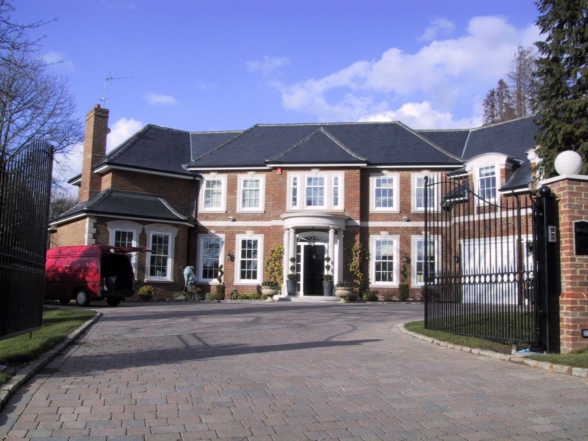 Large brick mansion with slate roof, white-trimmed windows, gated driveway, and red delivery van