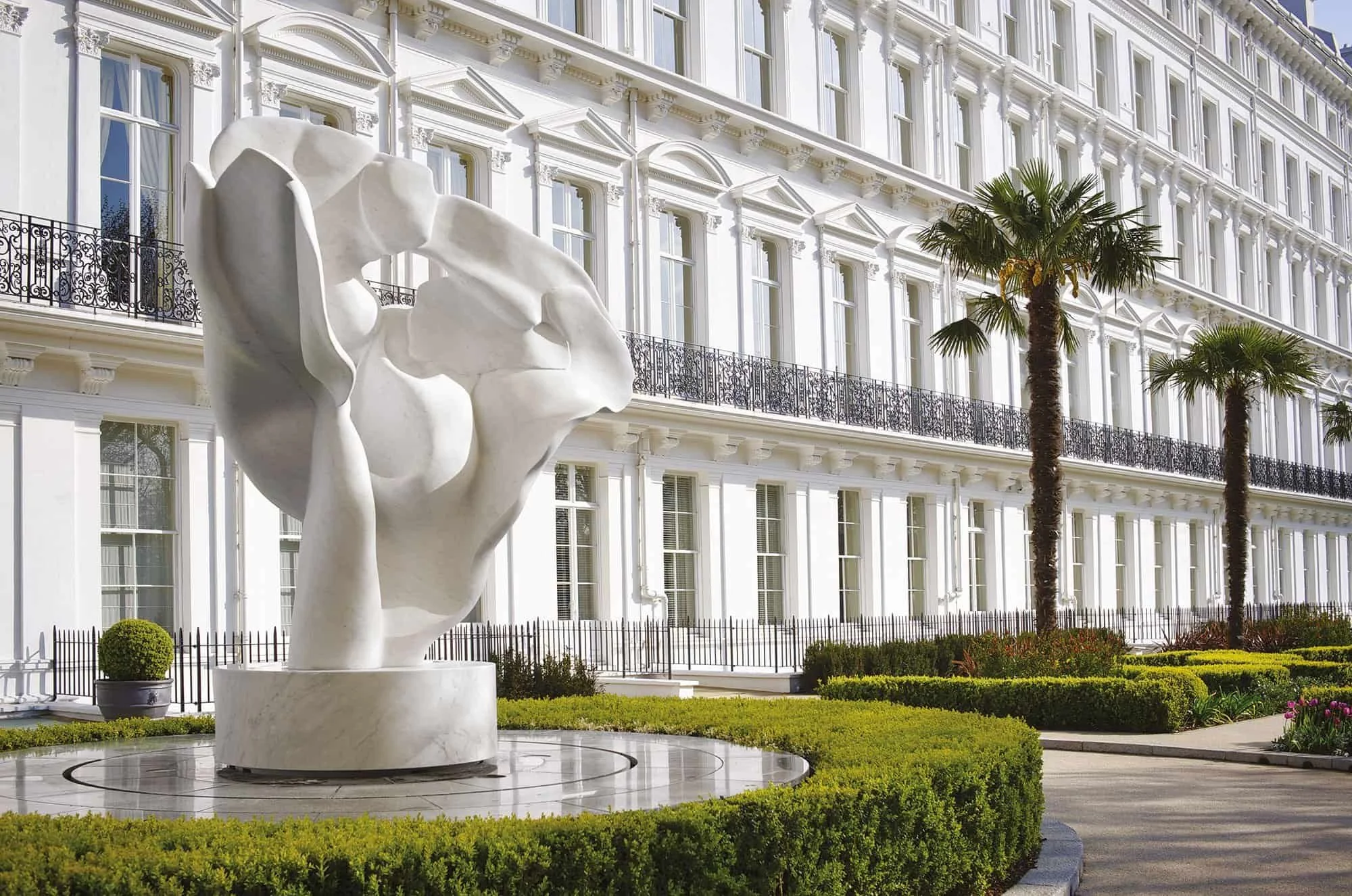 Abstract white sculpture in a manicured garden outside an ornate white townhouse with palm trees.
