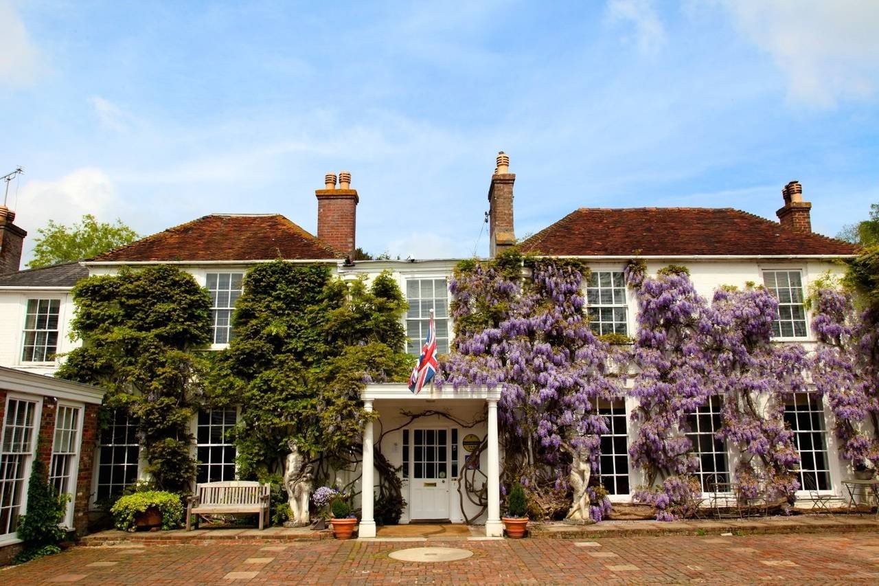 Historic English country house with red tile roofs, brick chimneys, lush gardens, flowering purple wisteria, and a Union Jack flag.