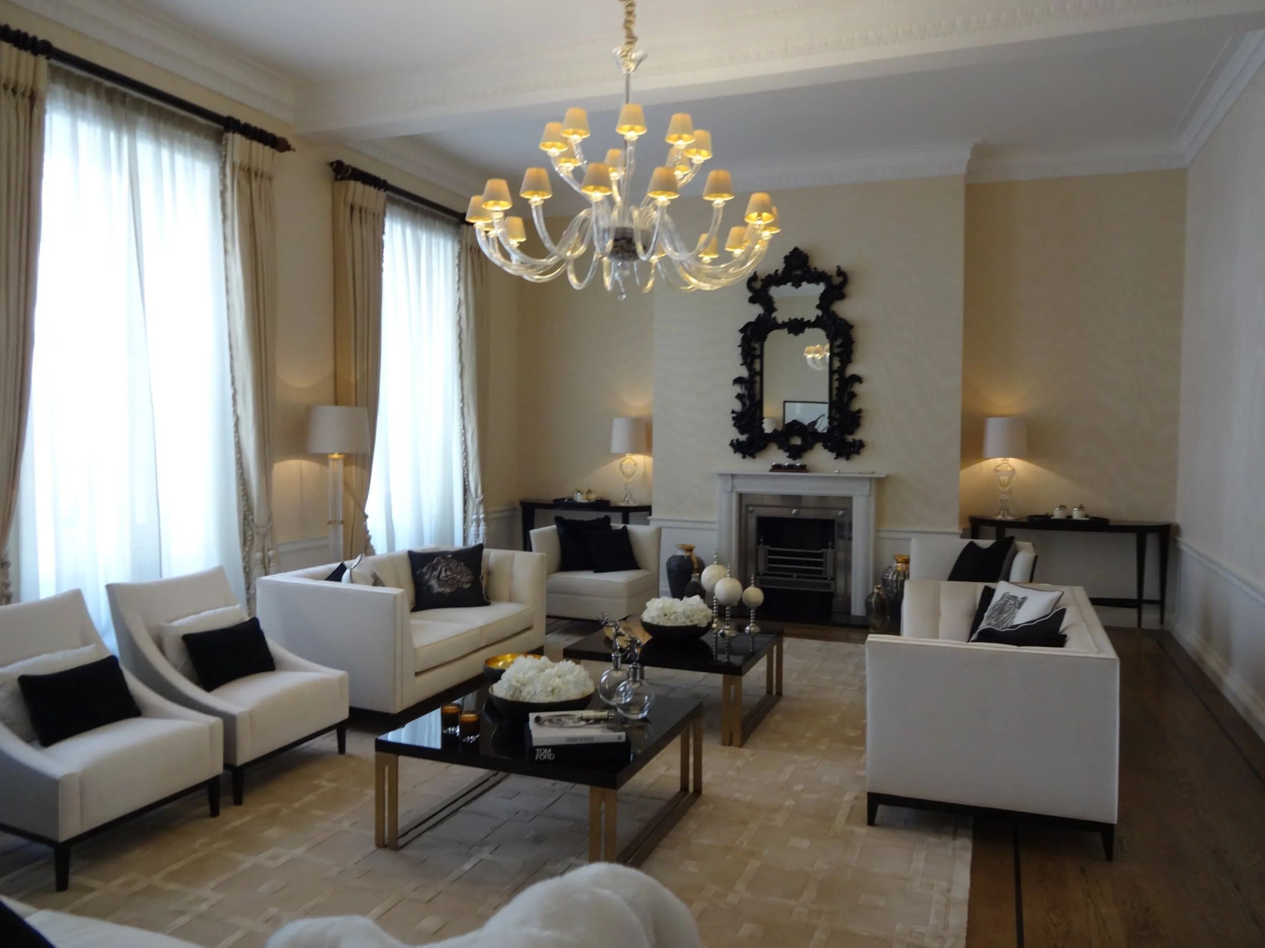 Elegant living room with white armchairs, black accents, chandelier, fireplace, ornate mirror, and coffee tables