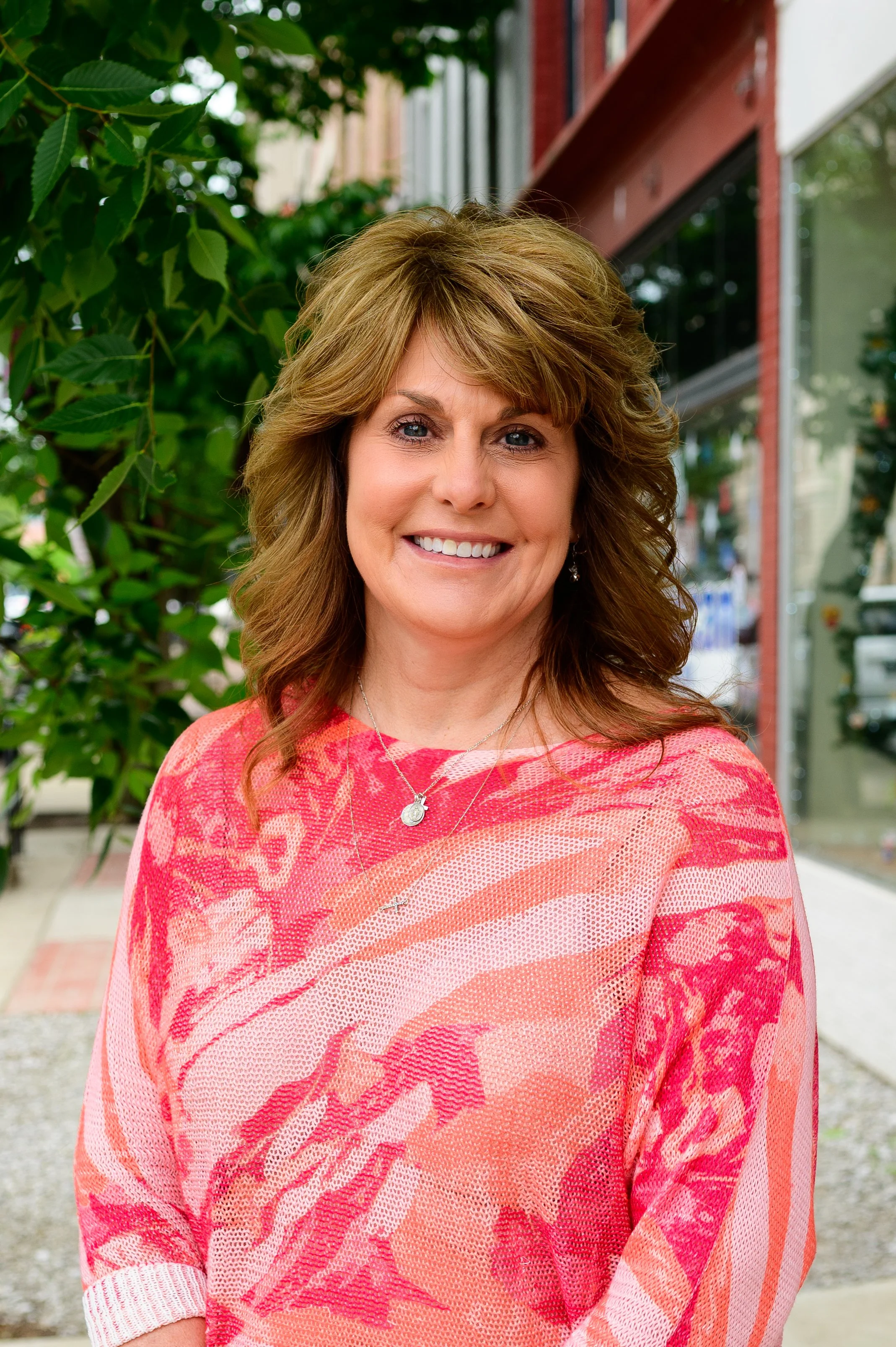 A woman with shoulder-length wavy light brown hair smiling, wearing a white denim jacket over a black and white patterned top, with layered necklaces, against a green background.
