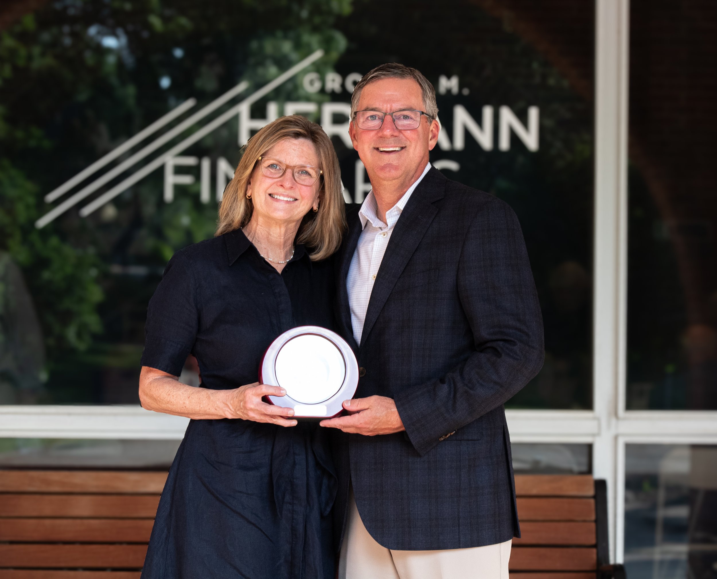 A smiling middle-aged woman holding a plaque, standing next to a smiling middle-aged man in a suit, both outdoors in front of a window with text, appearing to be at an award or recognition event.