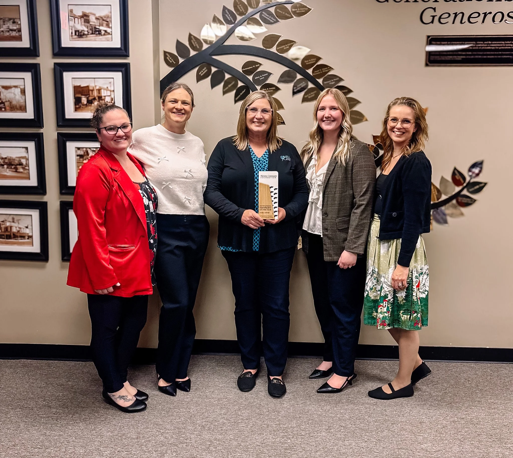 Five women standing in a row indoors, smiling, with a wall display of photographs and a large decorative tree behind them. The woman in the middle is holding a wooden award.