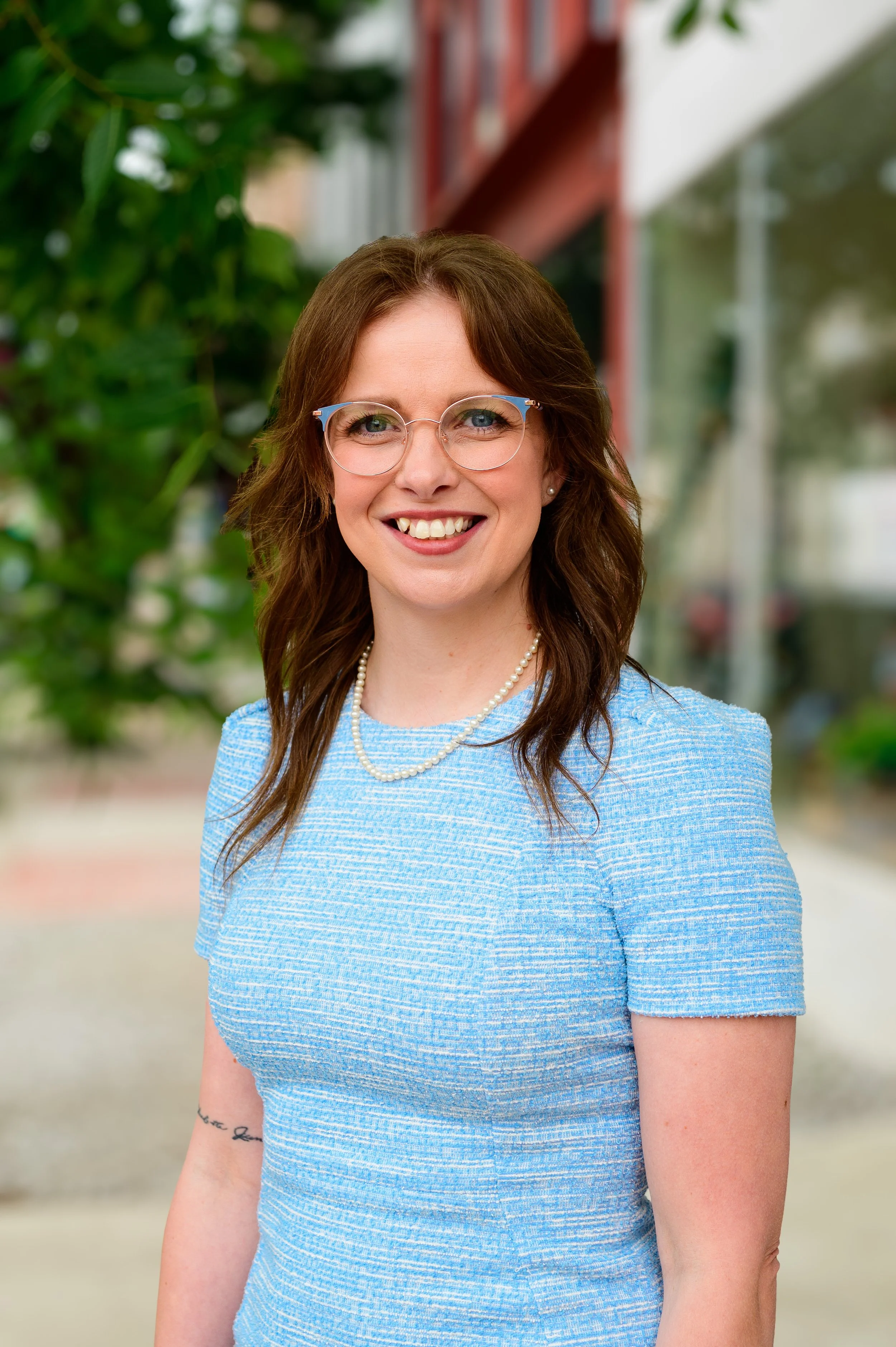 Portrait of a woman with glasses, smiling, wearing a beige blazer and jewelry, standing against a plain green background.