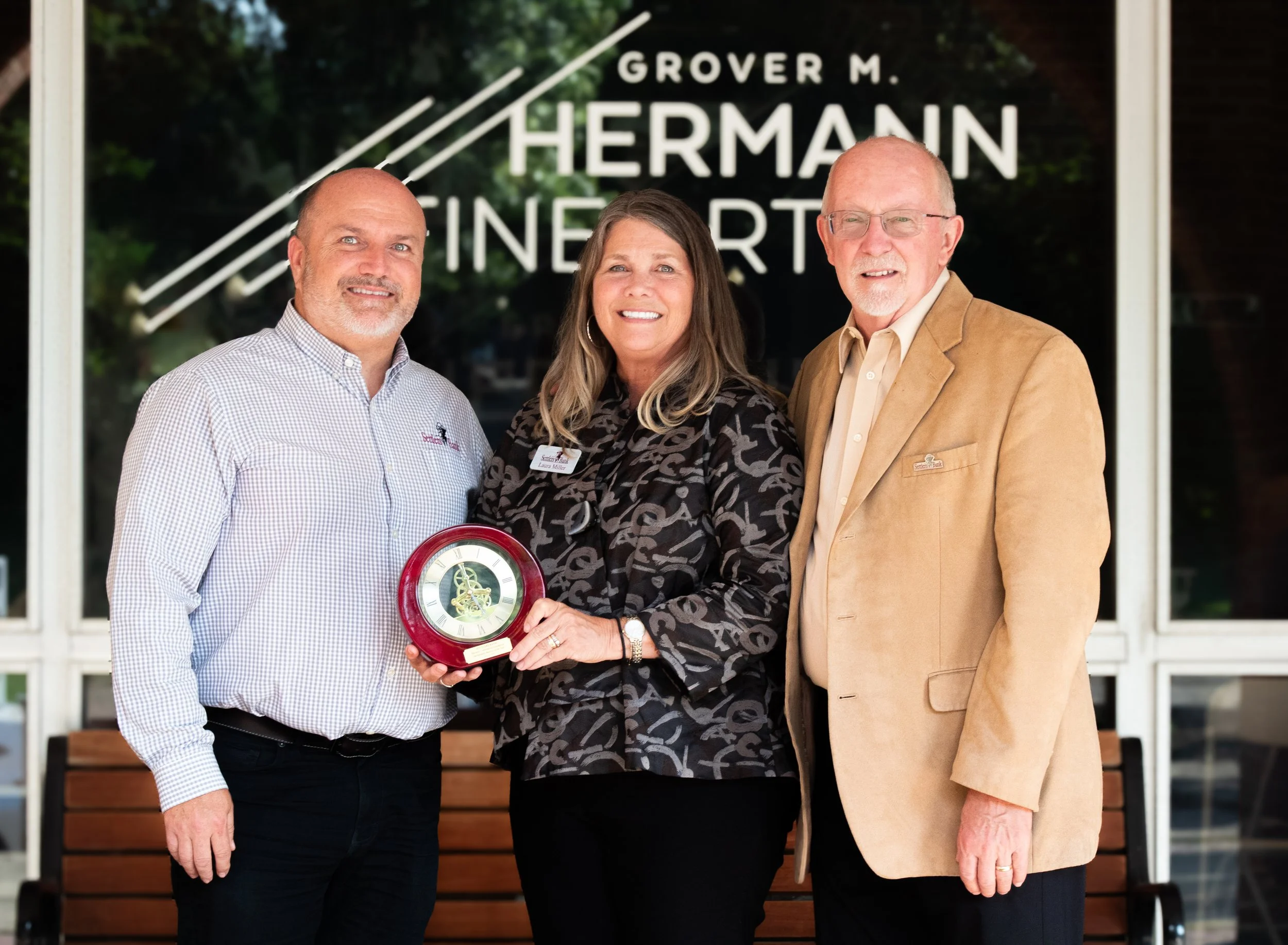 Three people standing outdoors in front of a building with a sign that reads 'GROVER M. HERMAN FINERTY.' The woman in the middle is holding a red and gold clock and is smiling. The two men on either side are also smiling, and the man on the right is wearing a tan blazer.