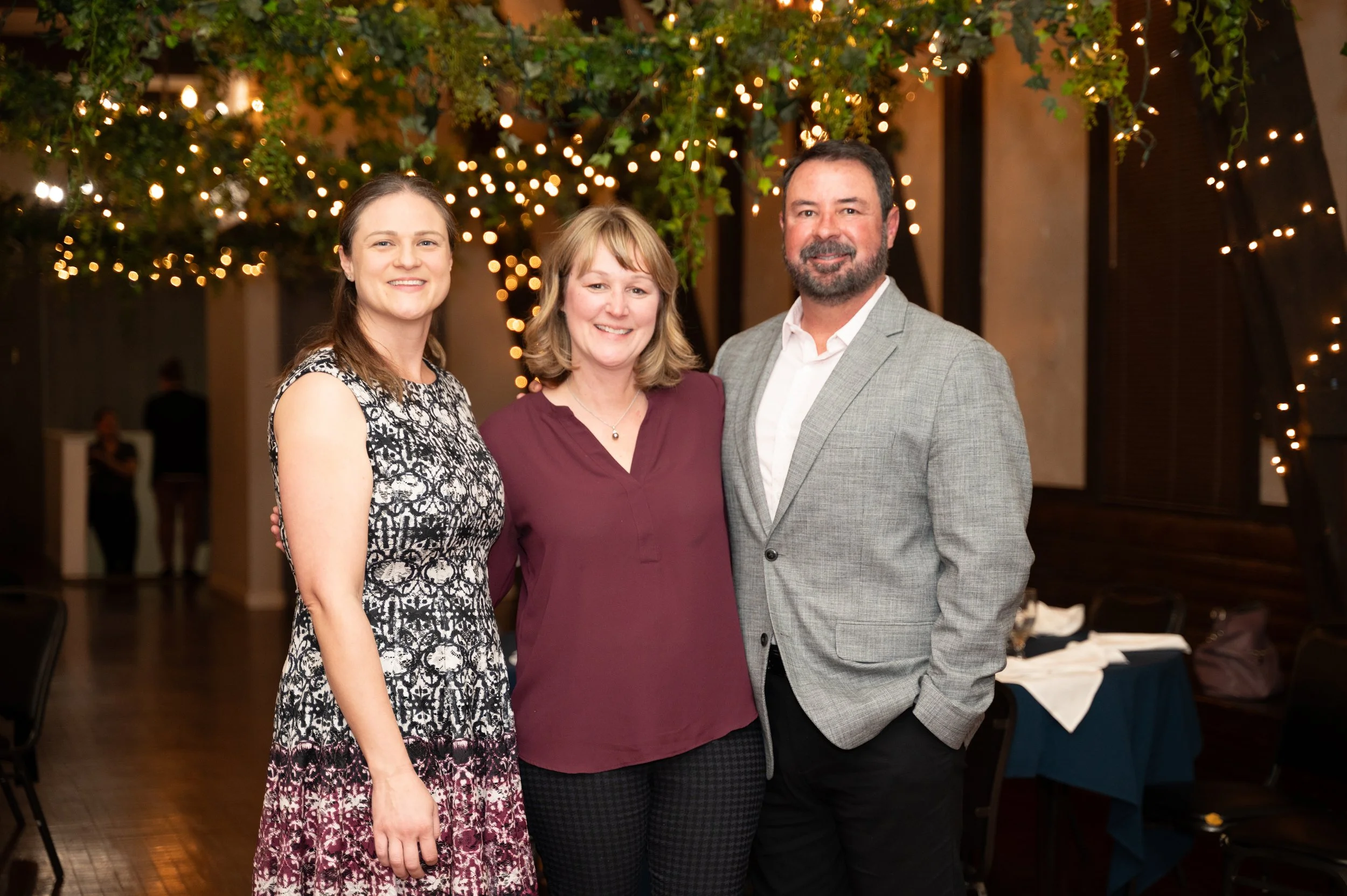 Three people standing together at a social event, smiling, in a dimly lit venue with string lights and greenery above them.