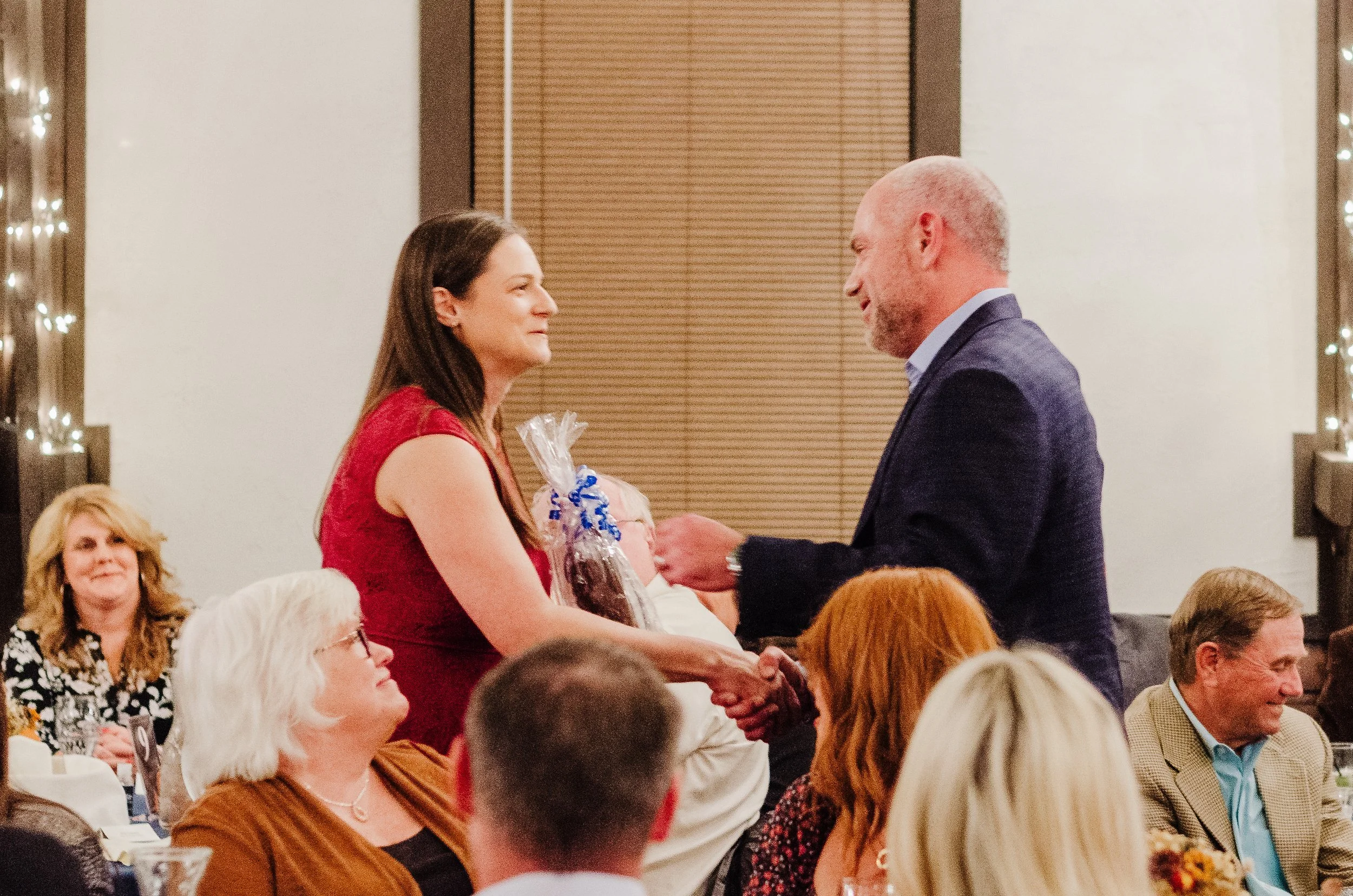 A woman in a red dress receives a gift and handshake from a man in a dark suit at a formal event, with seated guests watching.