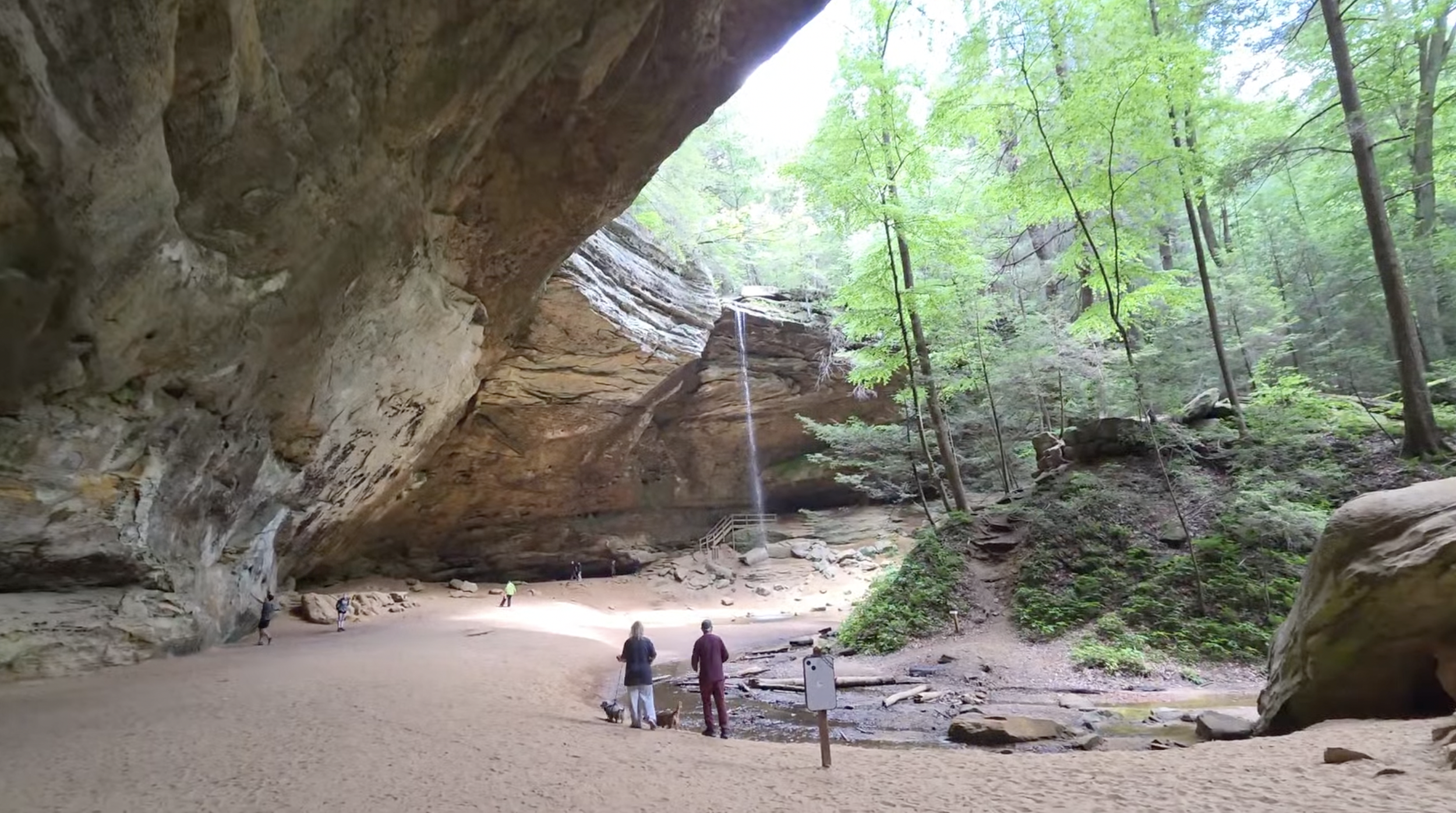 A small waterfall surrounded by a horseshoe shaped cliff with people around the bottom of the waterfall