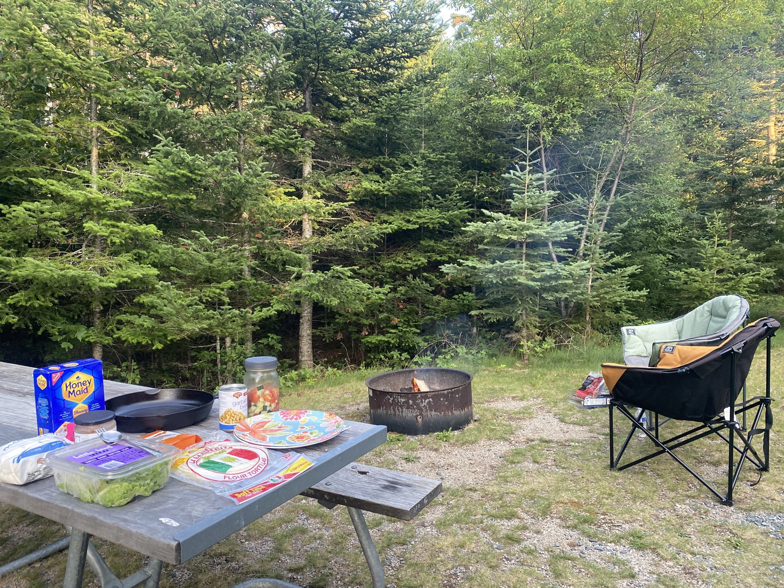 Forested campsite with campfire going, two camp chairs, and a table with food on top.