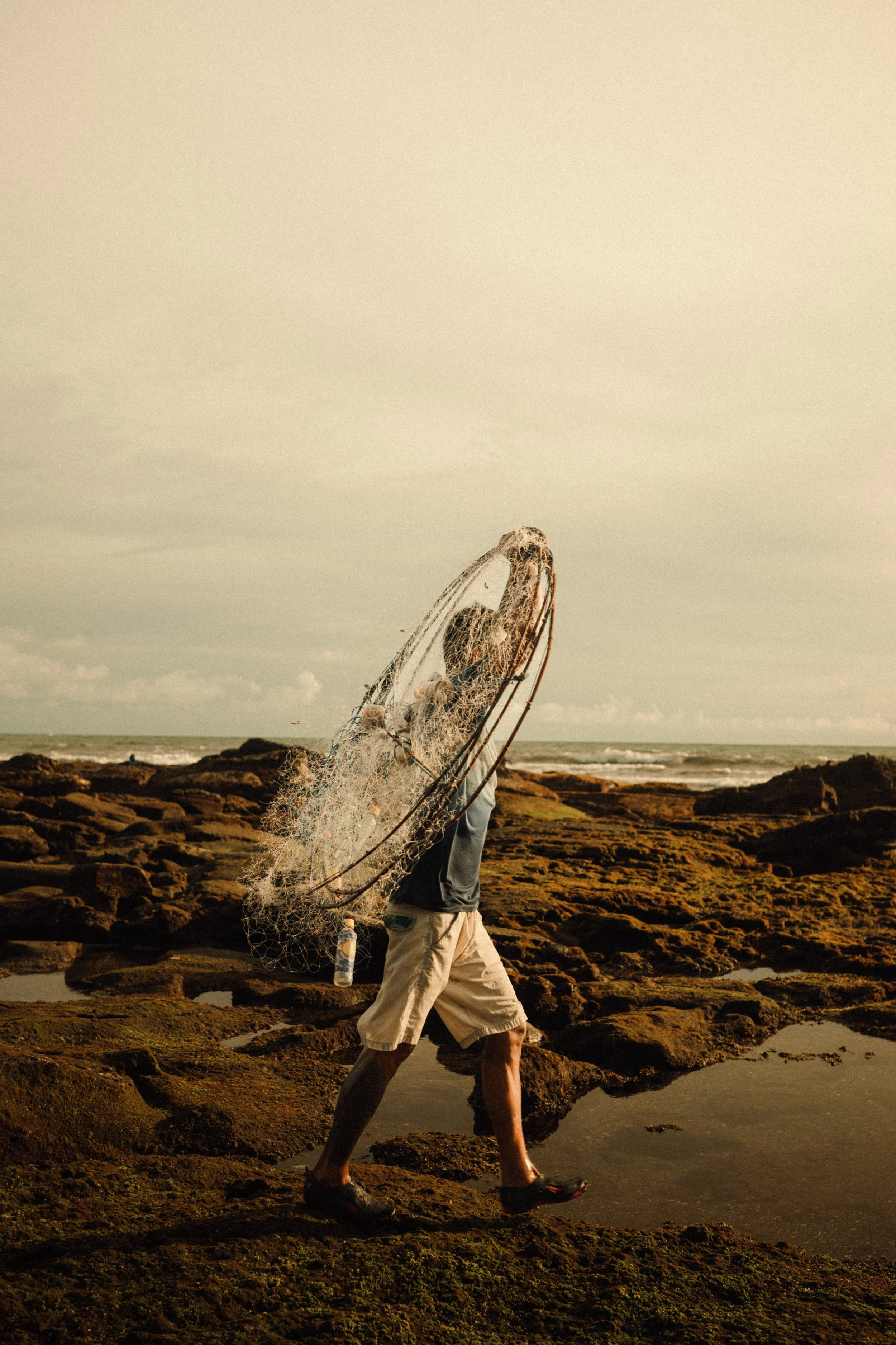 Uomo con pantaloncini chiari e maglietta scura che cammina su rocce lungo la costa con reti da pesca in mano