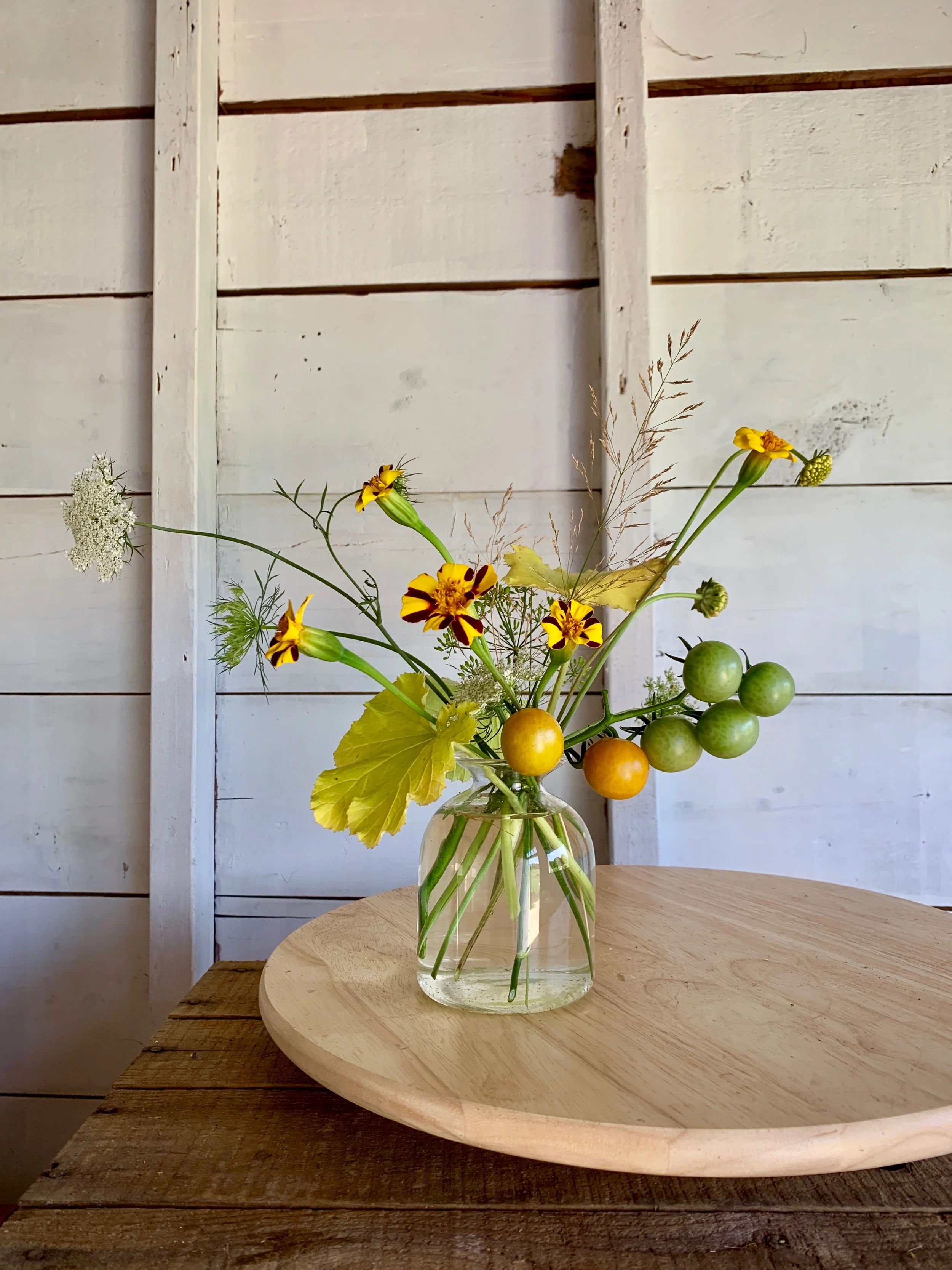 A clear glass vase with a bouquet of various wildflowers and green berries, placed on a round wooden board on a rustic wooden table against a white wooden wall background.