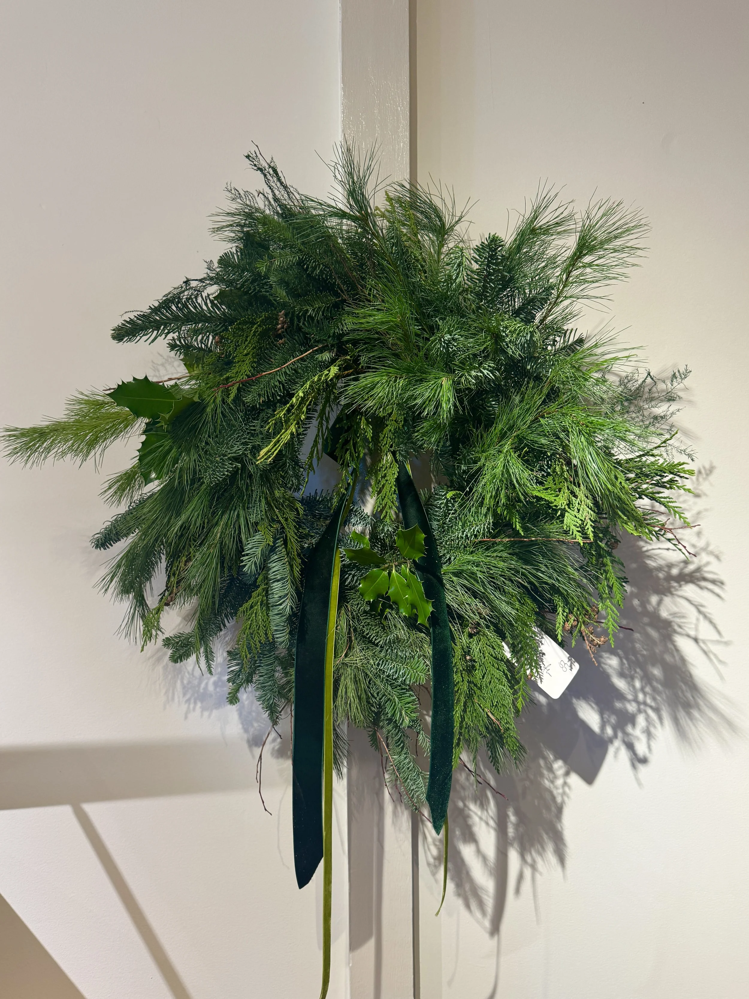 Green holiday wreath with various pine and holly foliage, hanging on a beige wall.