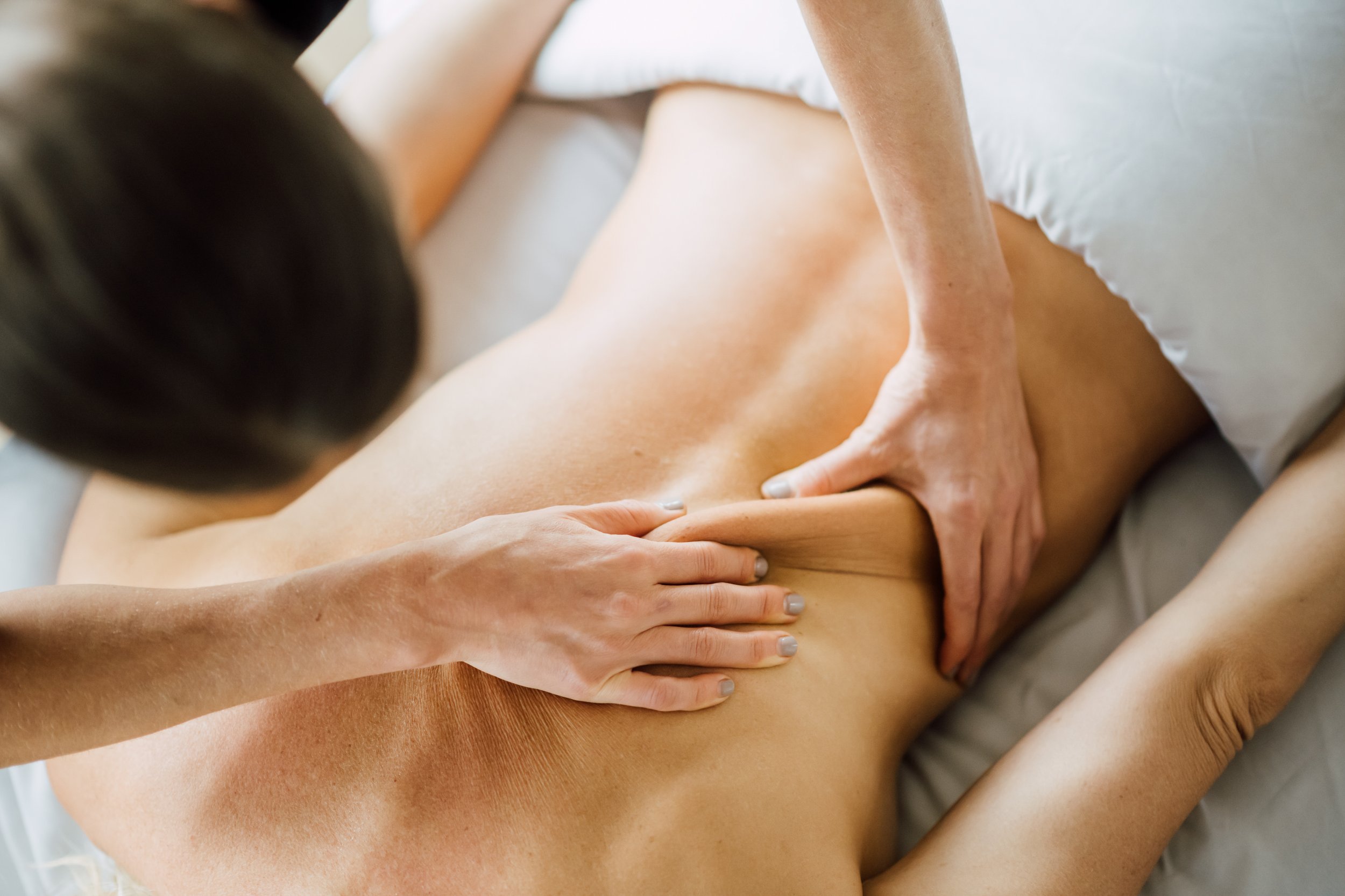 Massage therapist using both hands to apply firm pressure to a woman’s lower back while she lies face down on a treatment table.