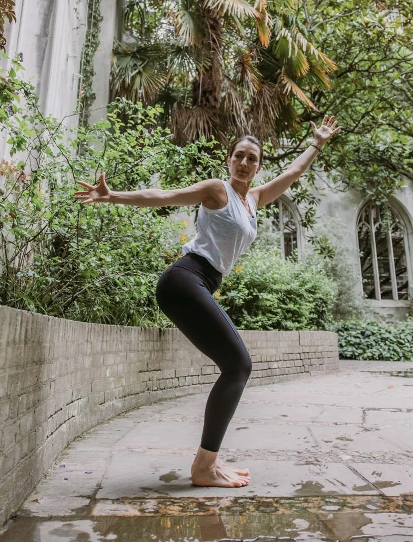 Sonia from Soniayogi practicing yoga outdoors in a lush garden with greenery and large palm trees, standing on a stone patio.