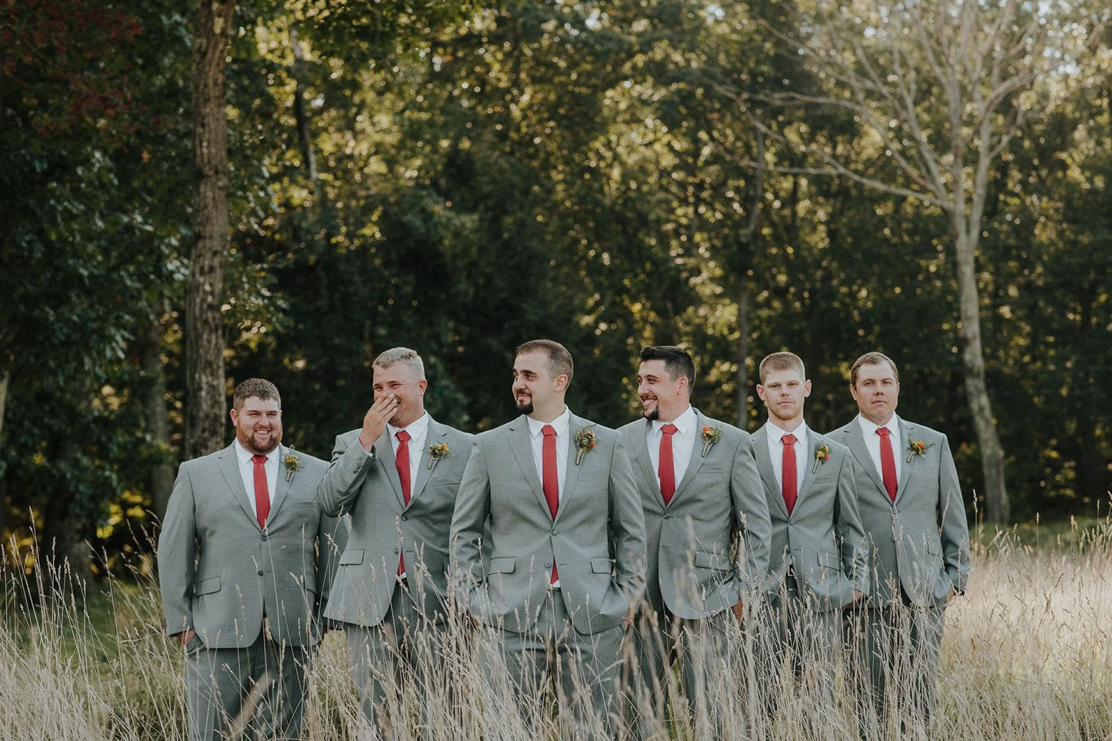 Seven men in gray suits with red ties walking through tall grass in a wooded outdoor setting, smiling and laughing.