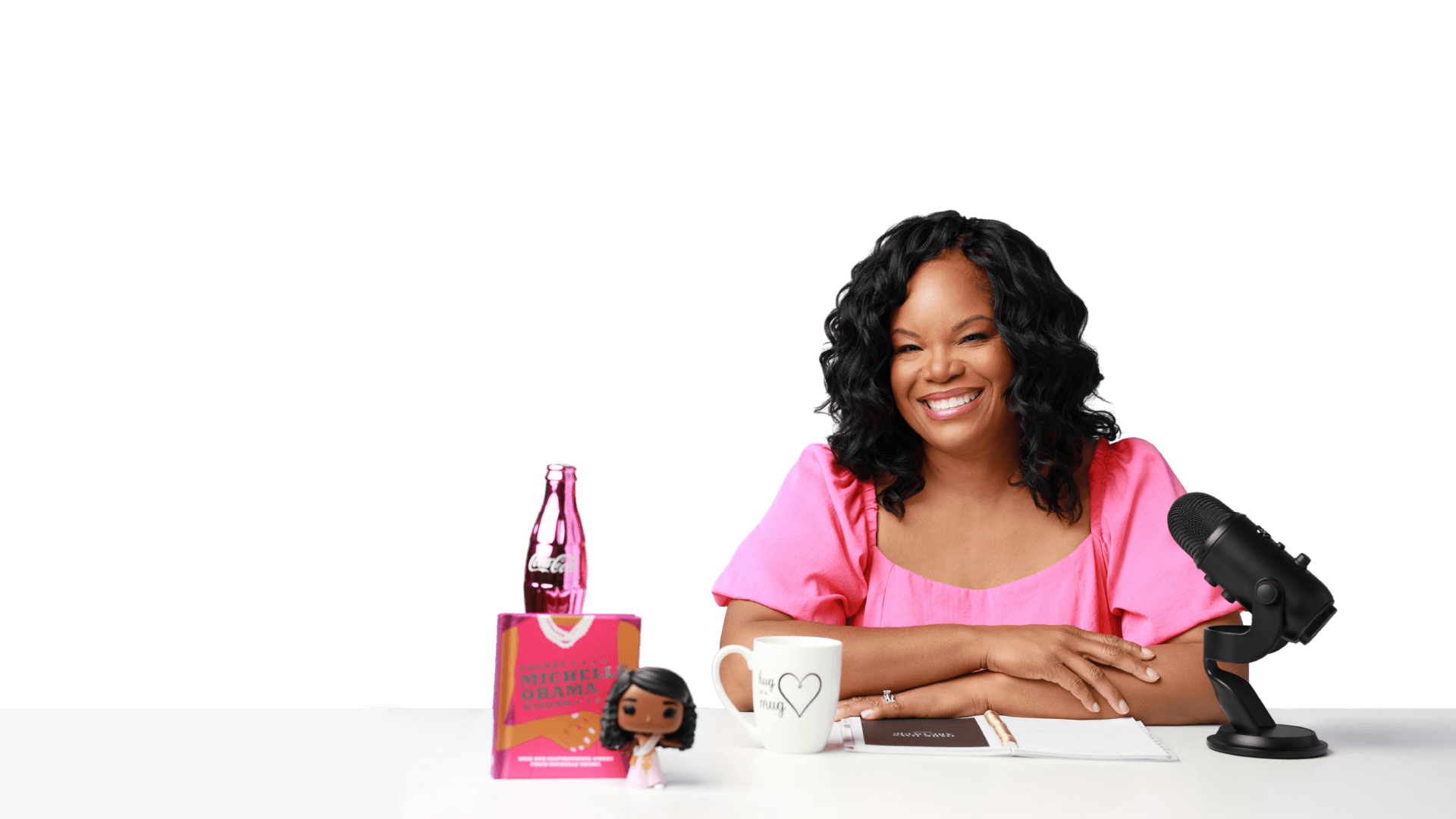 Nicole Gray smiling at a podcast desk in a pink top, with a microphone, mug, and notebook in front of her
