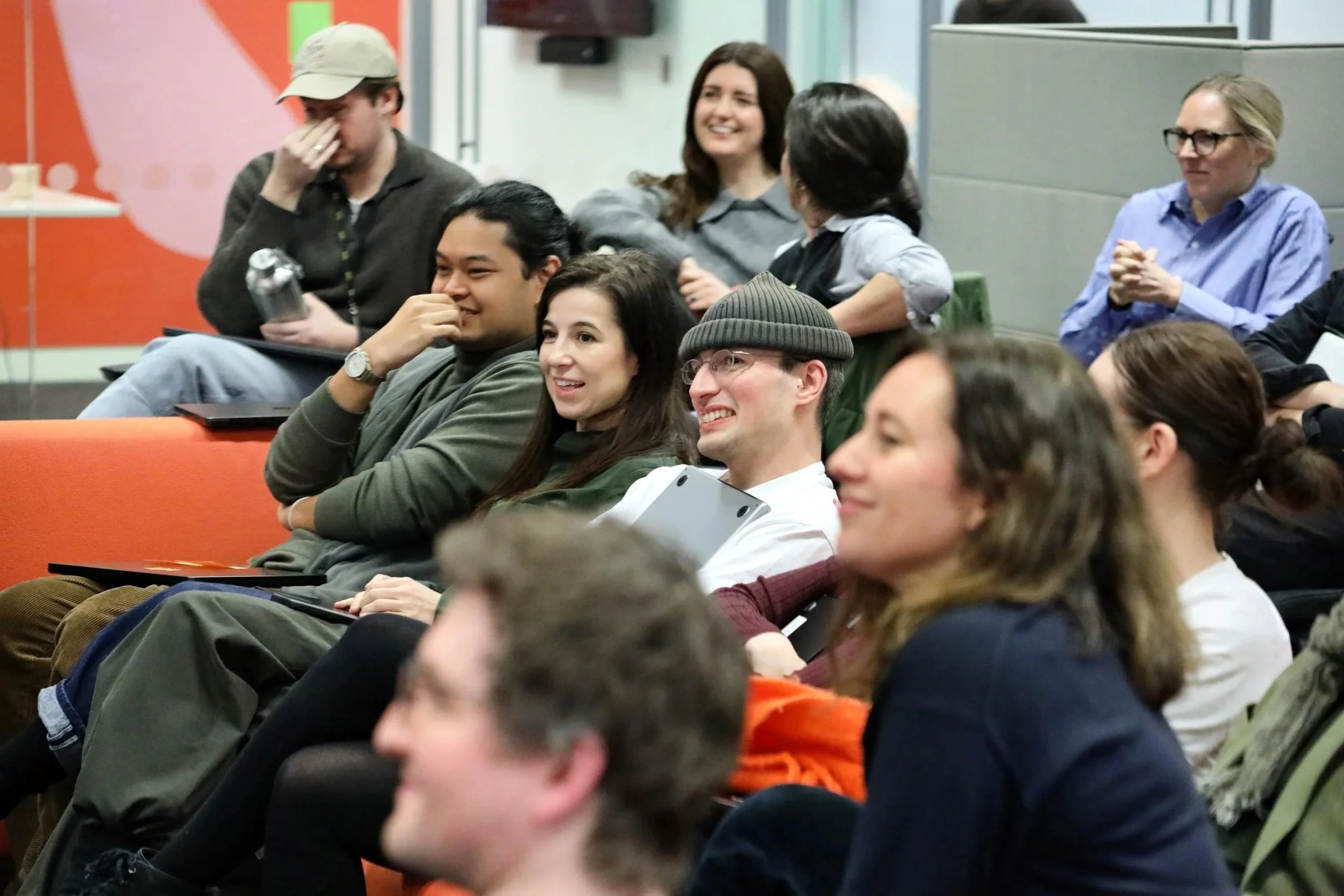 Group of diverse young adults attending a presentation or lecture, sitting in a classroom or conference room, some smiling, some laughing, engaged with the event.