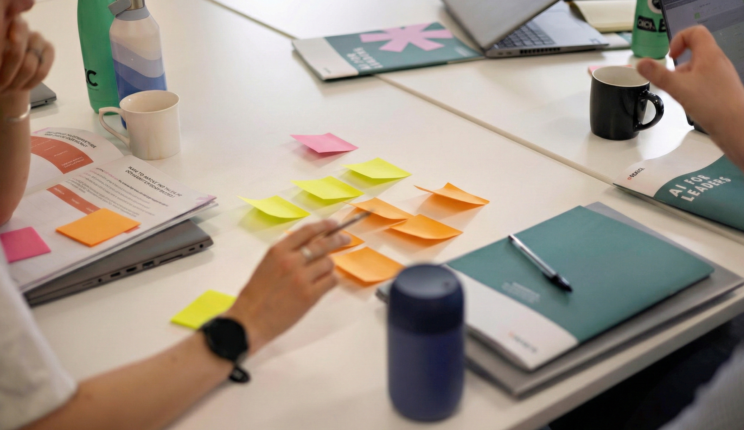 Meeting table with laptops, notebooks, coffee mugs, post-it notes, pens, water bottles, books, and people working or discussing.