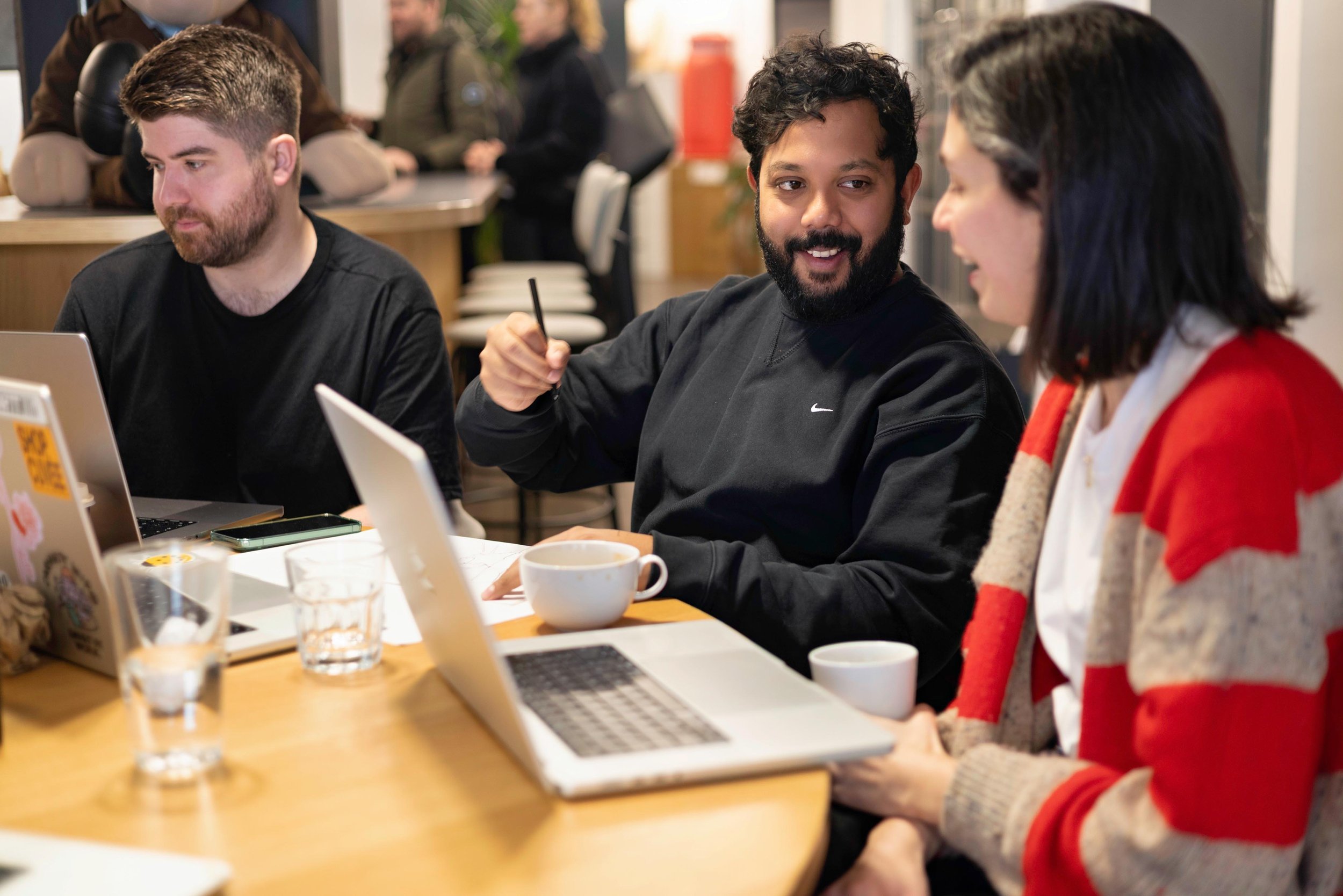 Three people sitting at a table in a cafe or coworking space. Two men and one woman are visible, engaged in conversation. The table has laptops, a mug, glasses, and other items. In the background, other people are present, blurred.