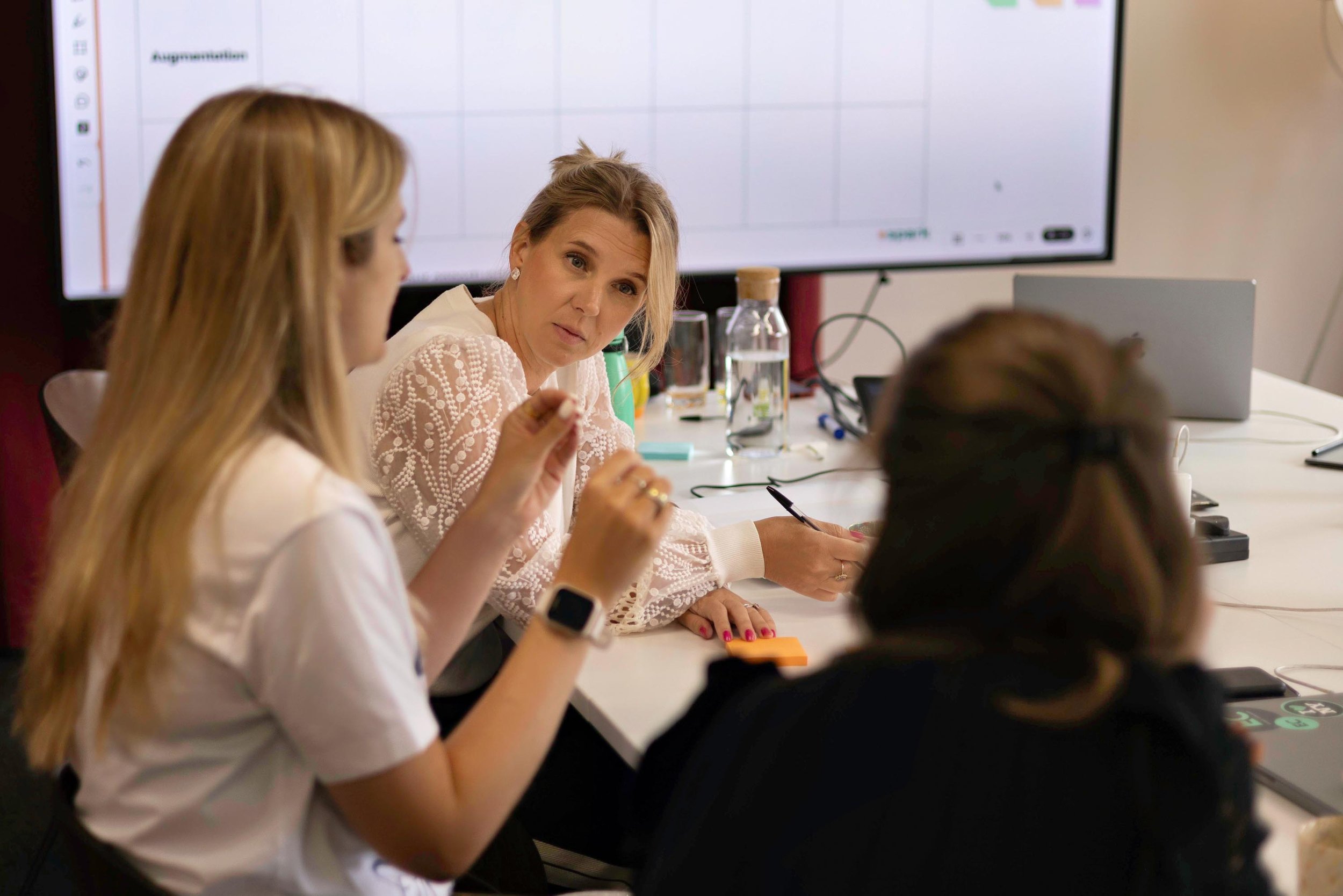 Group of women in a meeting room, engaged in discussion, with a large screen displaying a presentation in the background.