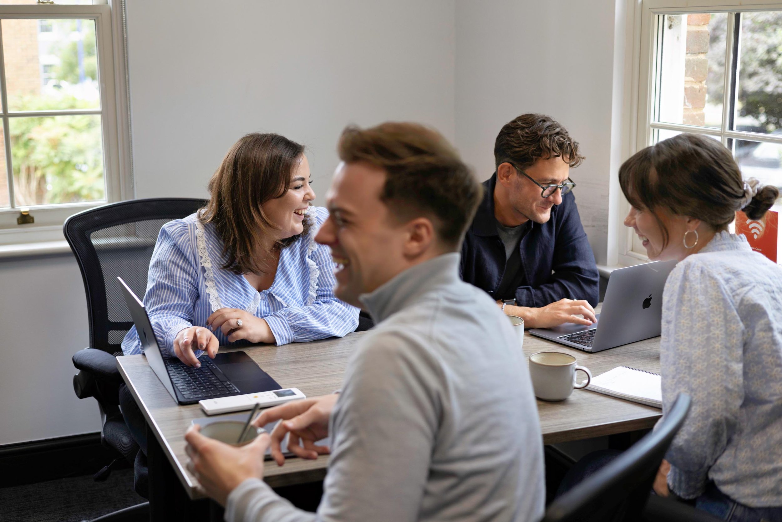 Group of five people sitting around a table in a bright office, working and chatting, with laptops and coffee mugs.