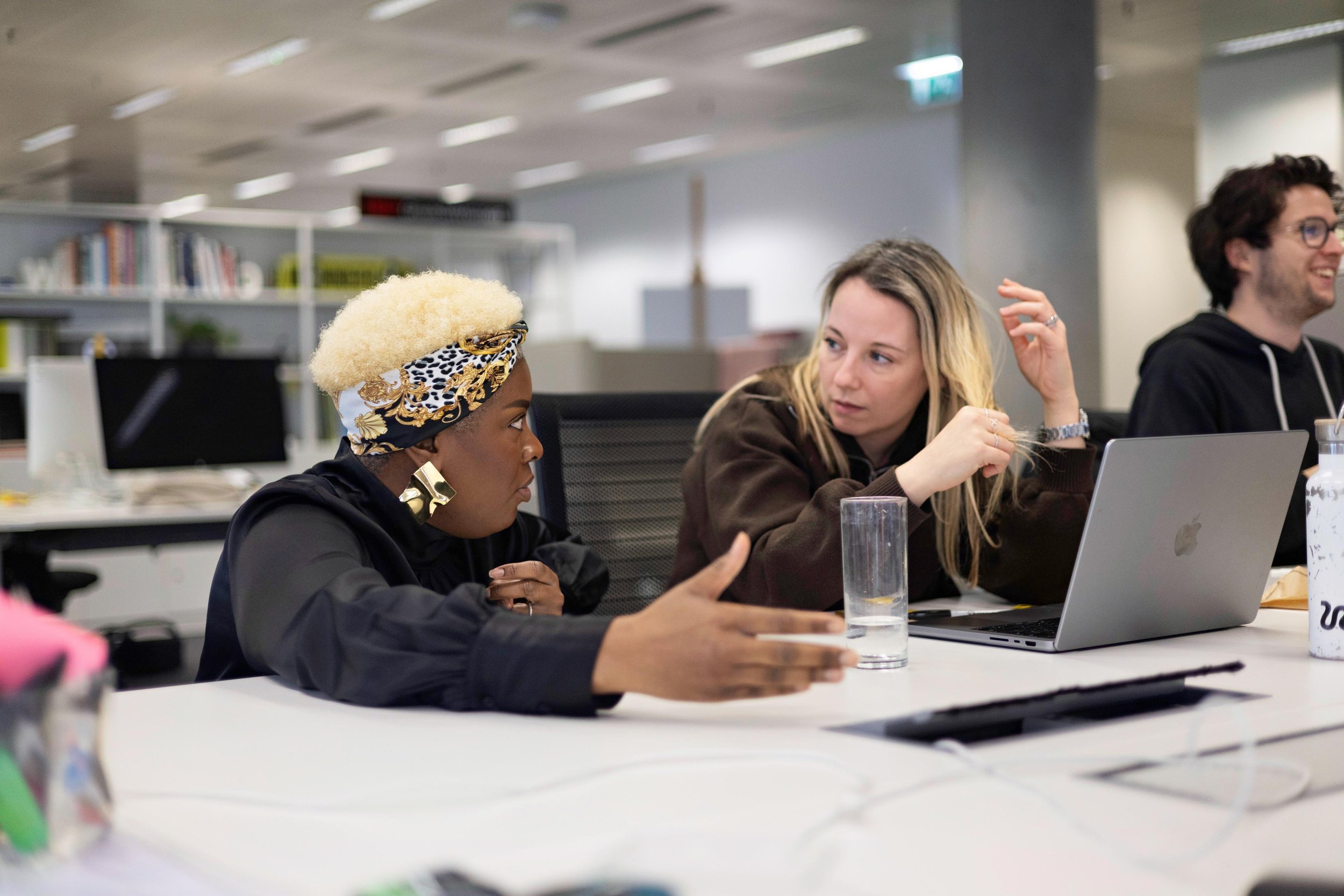 Three people having a discussion at a table in an office. The woman on the left has light skin, blonde hair, and is wearing a brown jacket. The woman in the middle has dark skin, blonde hair, and is wearing a black jacket with a large gold earring and a patterned headscarf. The man on the right has light skin, dark hair, glasses, and a black hoodie with white drawstrings. There is a laptop, a water glass, and some papers on the table.