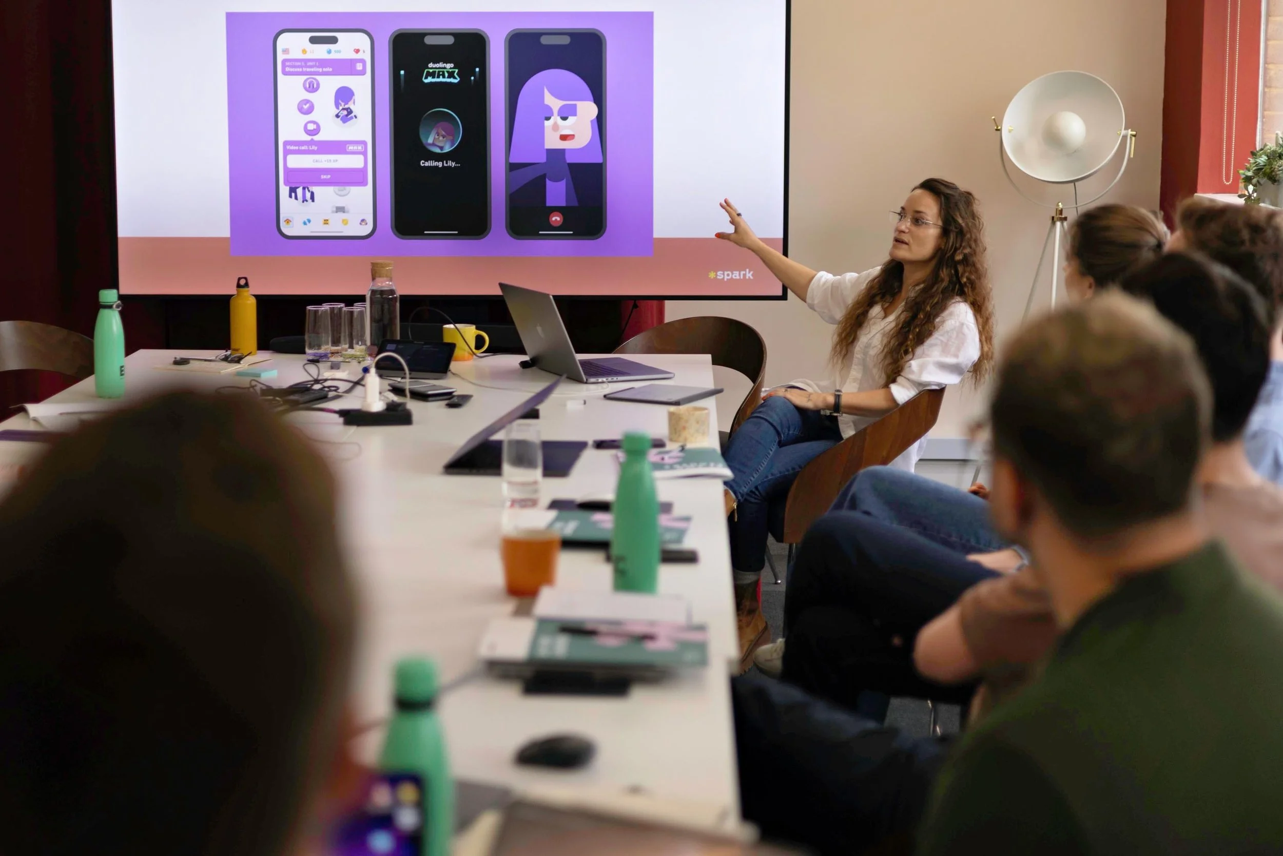 A woman is giving a presentation to a group of people in a conference room. She is pointing at a large screen displaying three smartphone screens with app interfaces. The room has a table with various items, including water bottles, a laptop, and notebooks. The audience is seated around the table, attentively listening.
