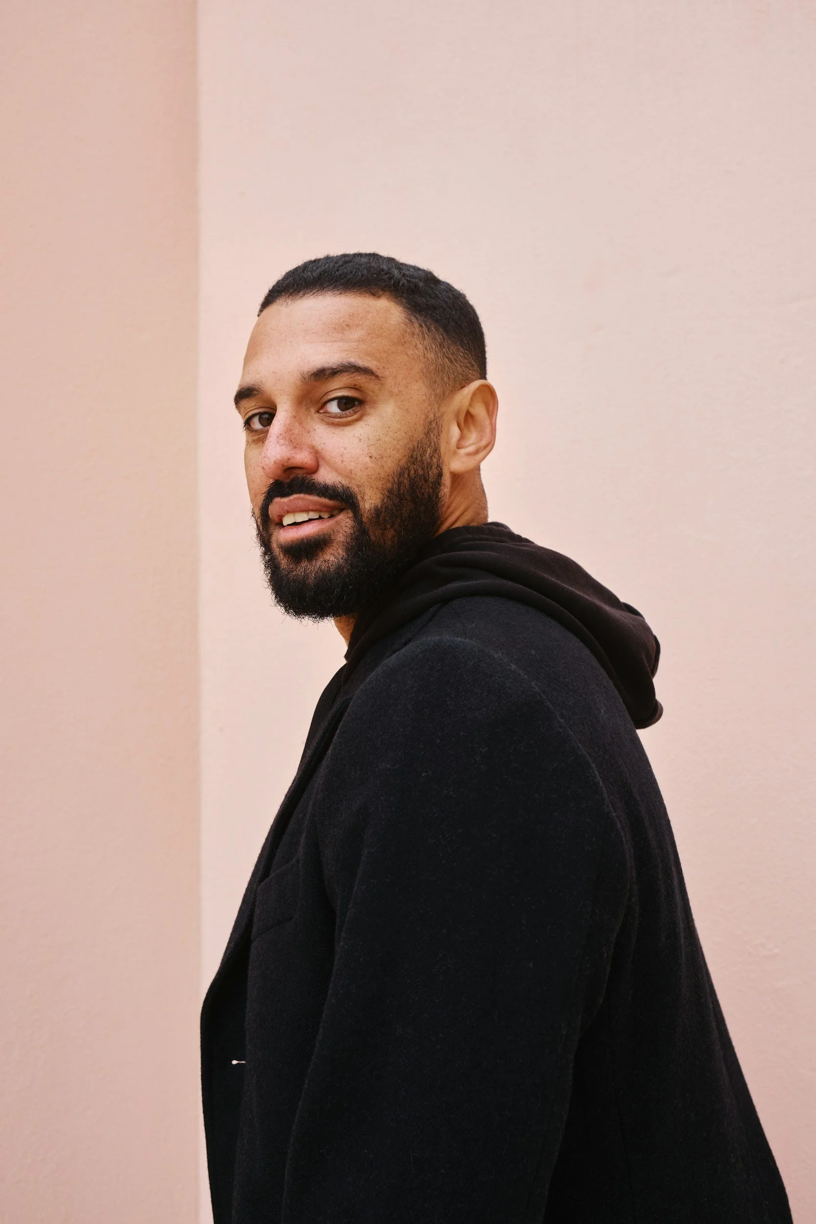A young man with short dark hair, a beard, and wearing a black hoodie, looking back over his shoulder against a plain pink wall.