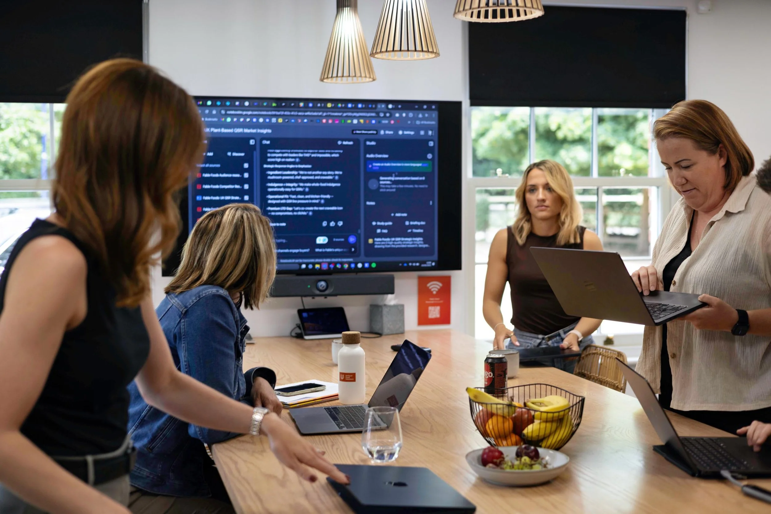 Group of women in a meeting room with laptops and a large screen displaying a social media dashboard, with a basket of fruit and bottles on the table.