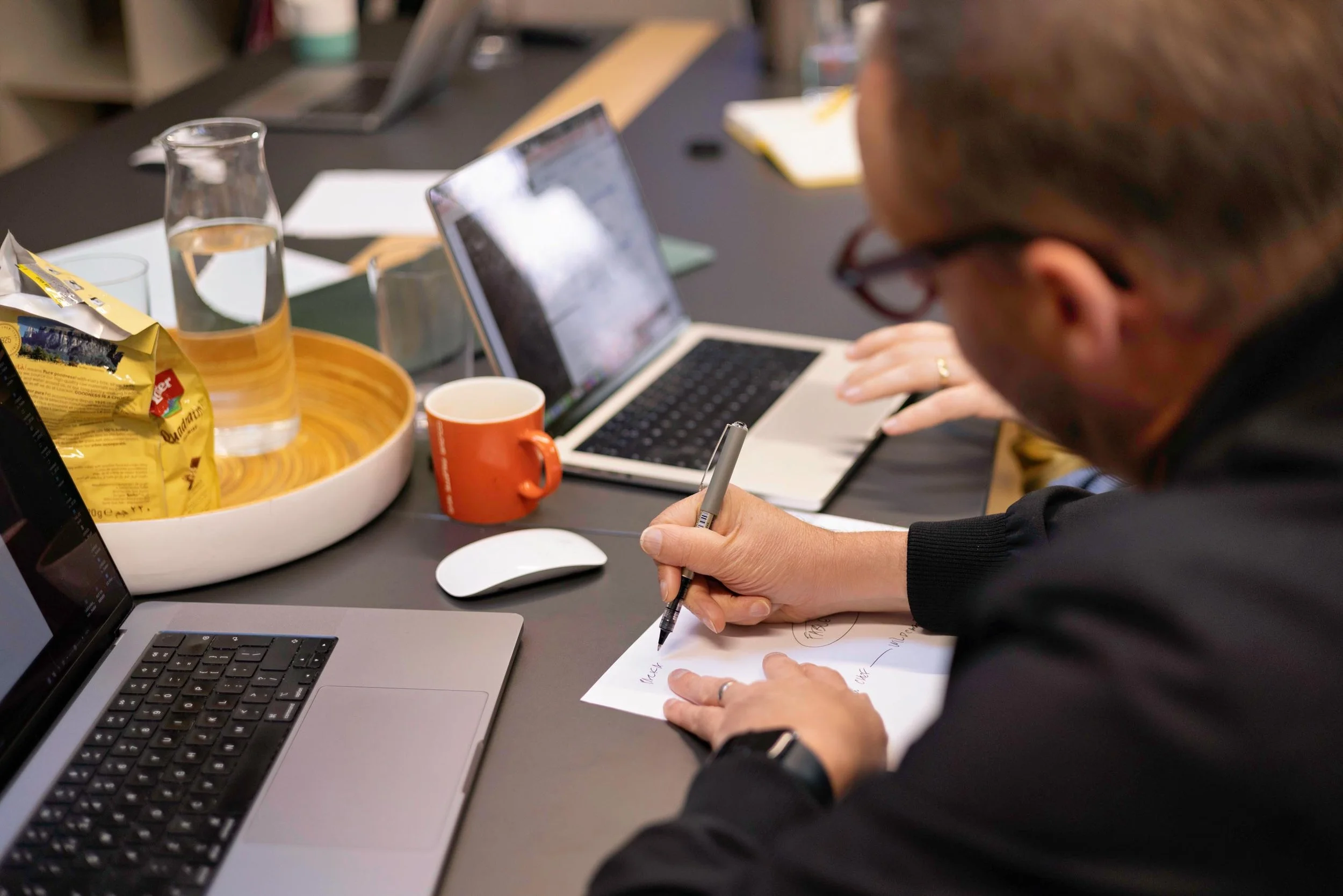 A person with brown hair and glasses writing on a piece of paper with a pen, surrounded by laptops, a tablet, a glass of water, a mug, and snacks on a dark desk.