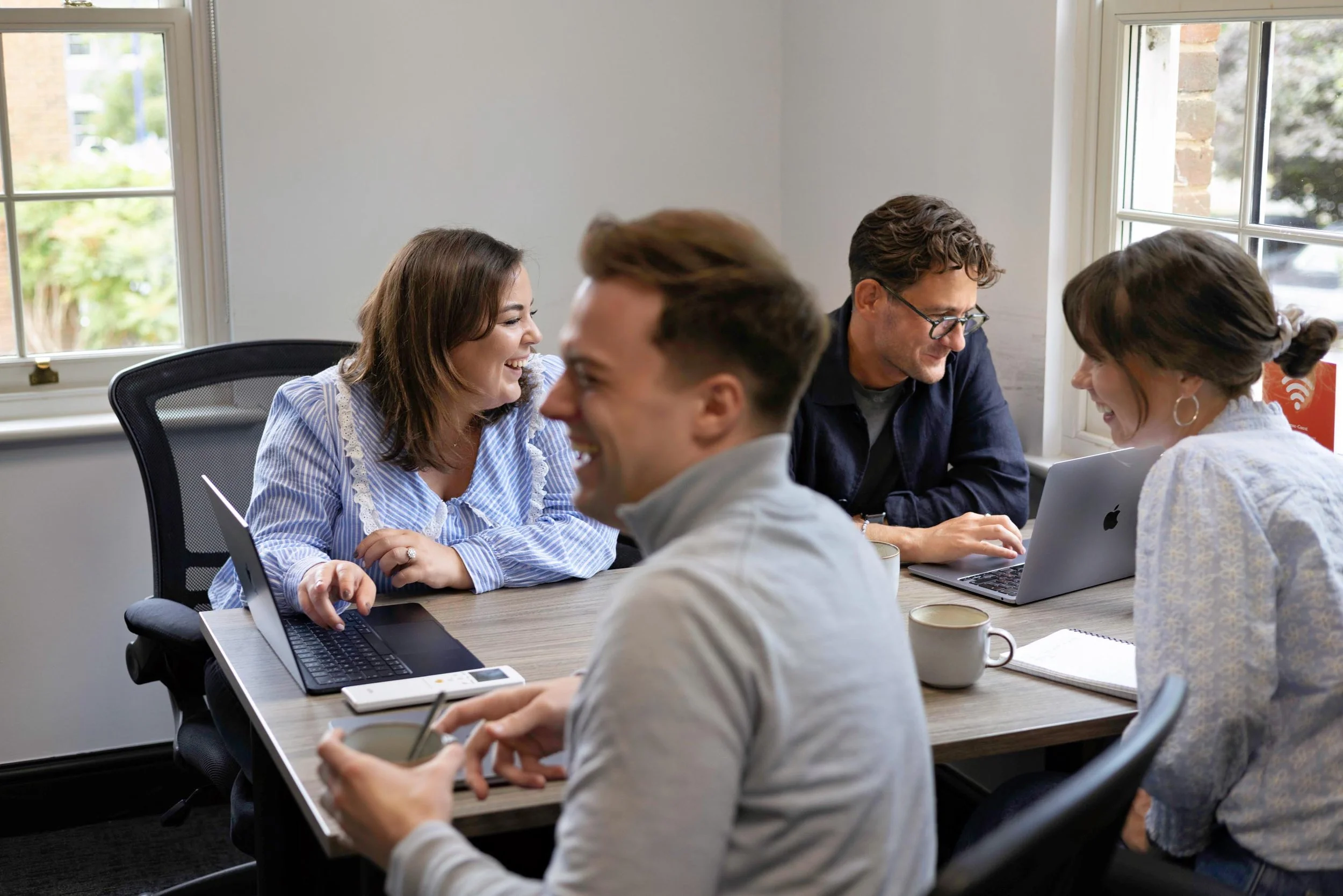 Group of five diverse people sitting around a conference table in a meeting room, smiling and engaged in conversation, with laptops, notebooks, and coffee cups on the table.