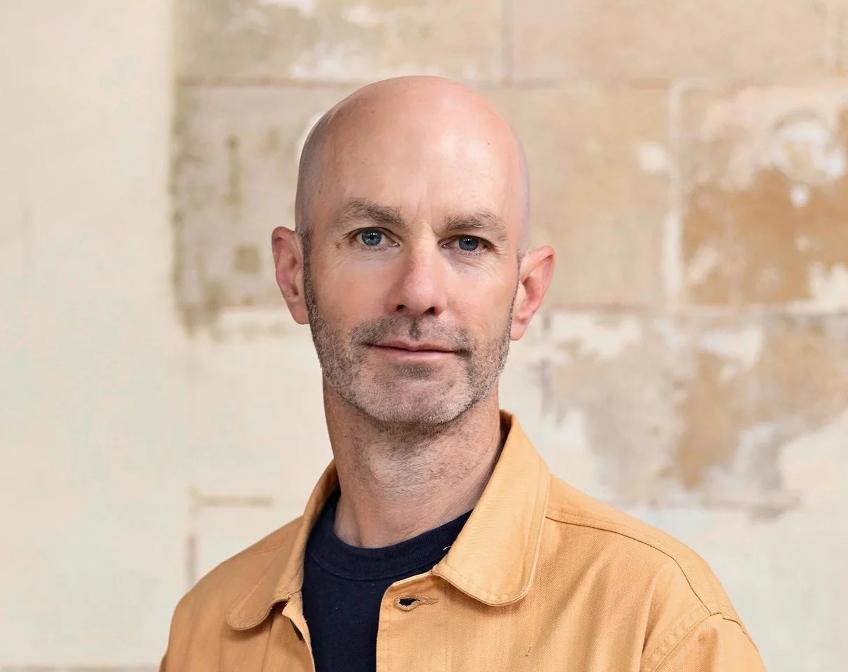 Portrait of a bald man with blue eyes, slight beard, wearing a beige shirt over a navy blue shirt, standing in front of a light-colored, textured background.