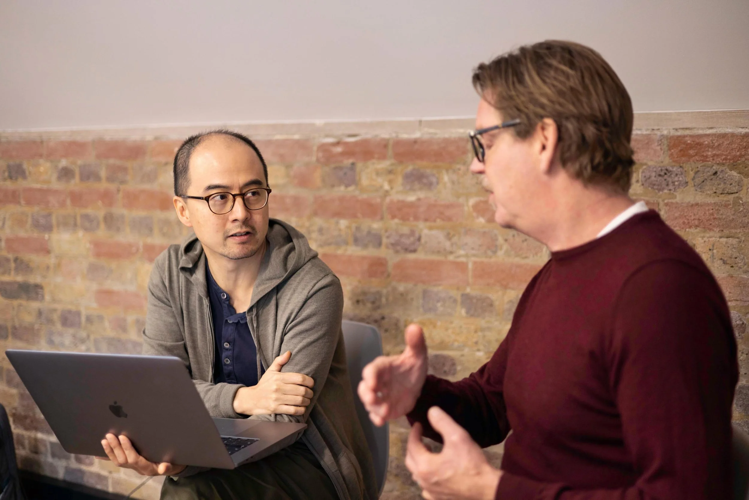 Two men engaged in a conversation indoors, sitting by a brick wall. One man, with glasses and wearing a grey hoodie, is looking attentively at the other man, who is wearing glasses and a maroon sweater, and appears to be explaining something.
