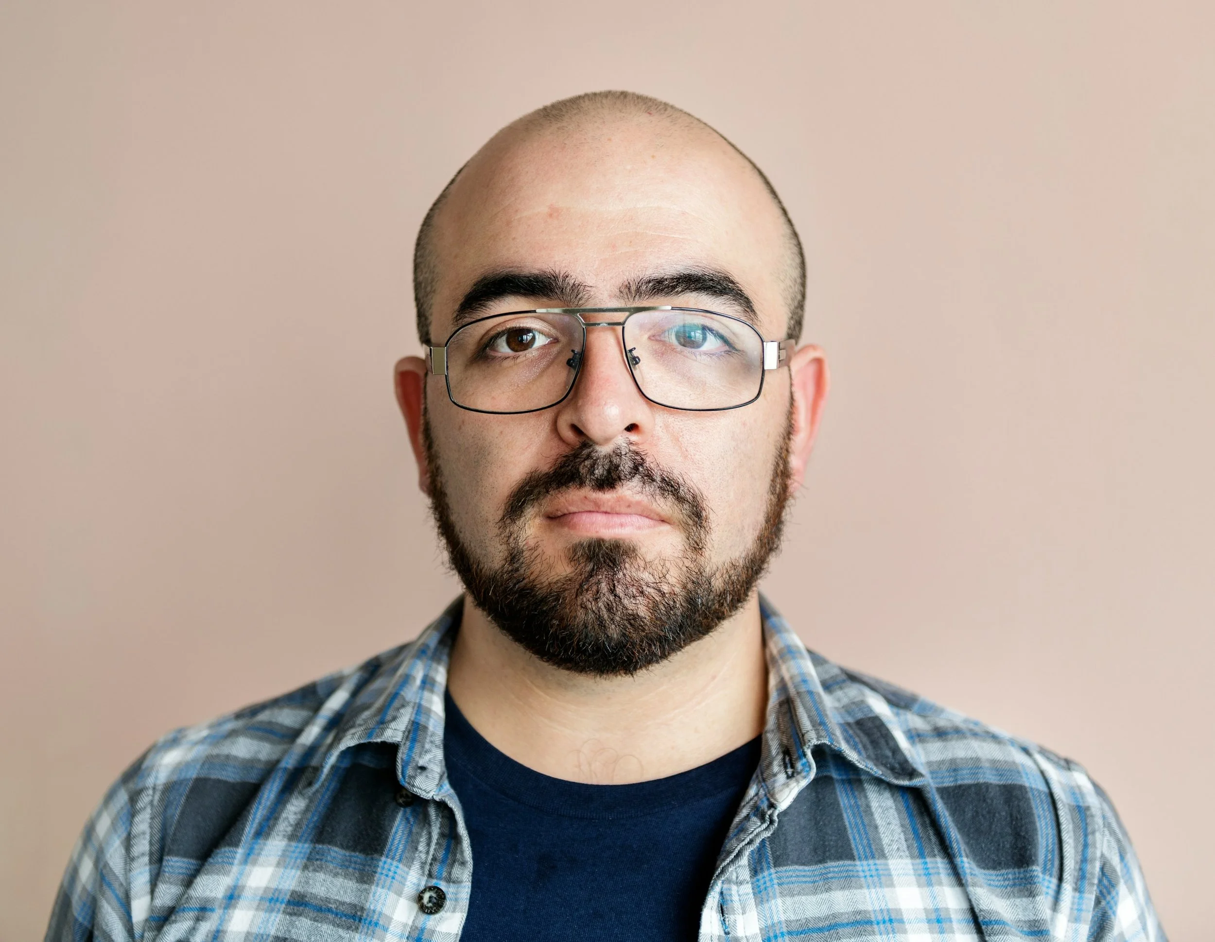 A man with a close-cropped beard and mustache wearing glasses and a blue plaid shirt, looking directly at the camera against a plain beige background.