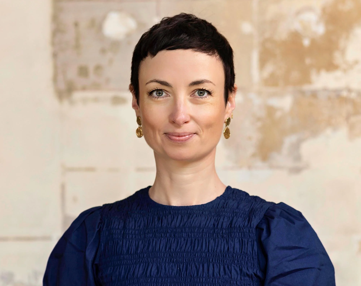 A woman with short dark hair wearing a navy blue top and gold earrings, standing in front of a textured beige wall.