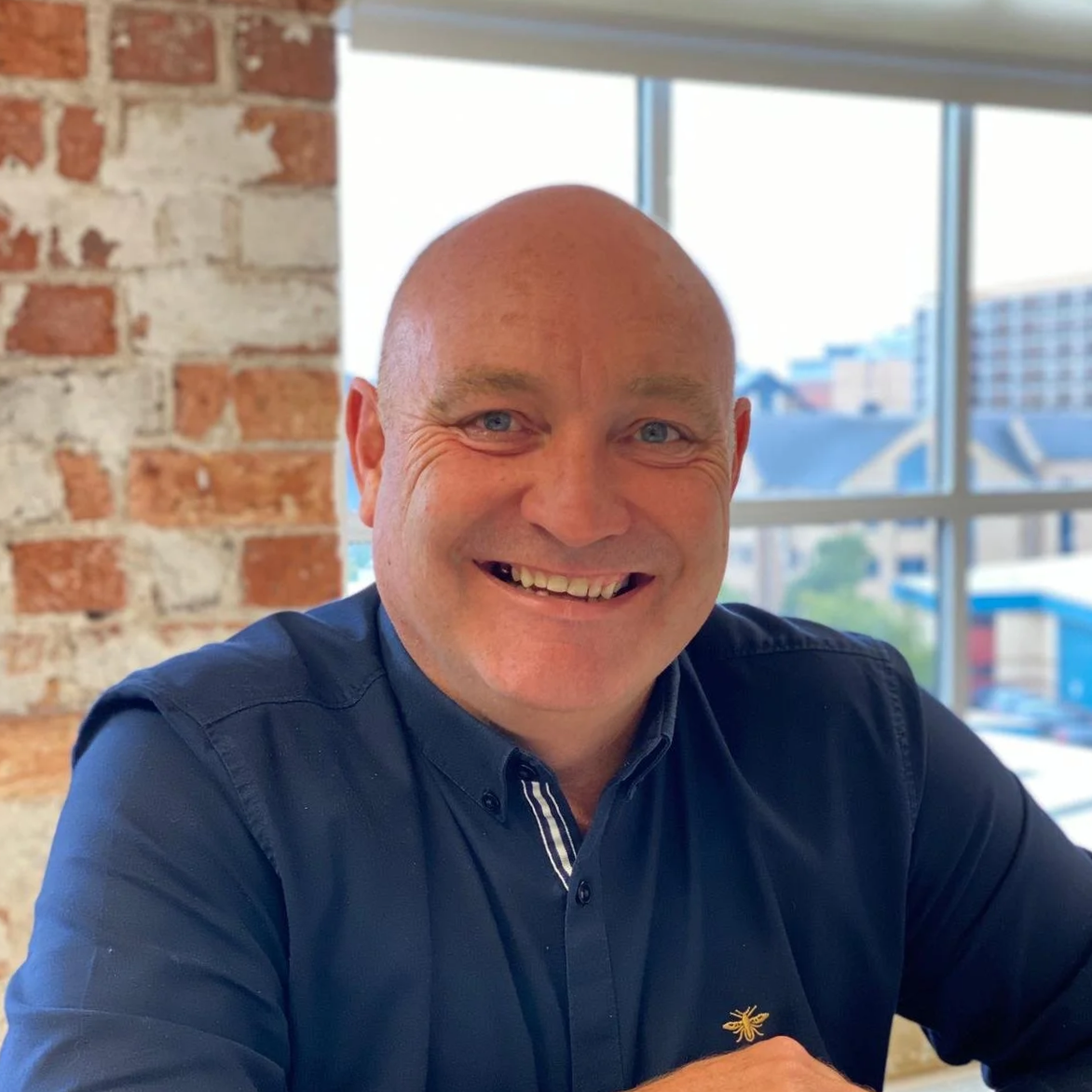 A smiling bald man wearing a navy blue shirt sitting indoors with a brick wall on the left and large windows showing buildings outside in the background.
