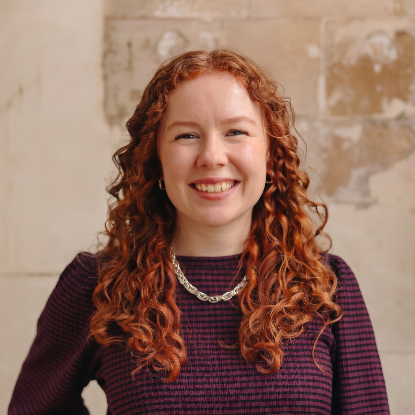 A young woman with curly red hair, glasses, and a patterned shirt, smiling in front of a textured beige wall.