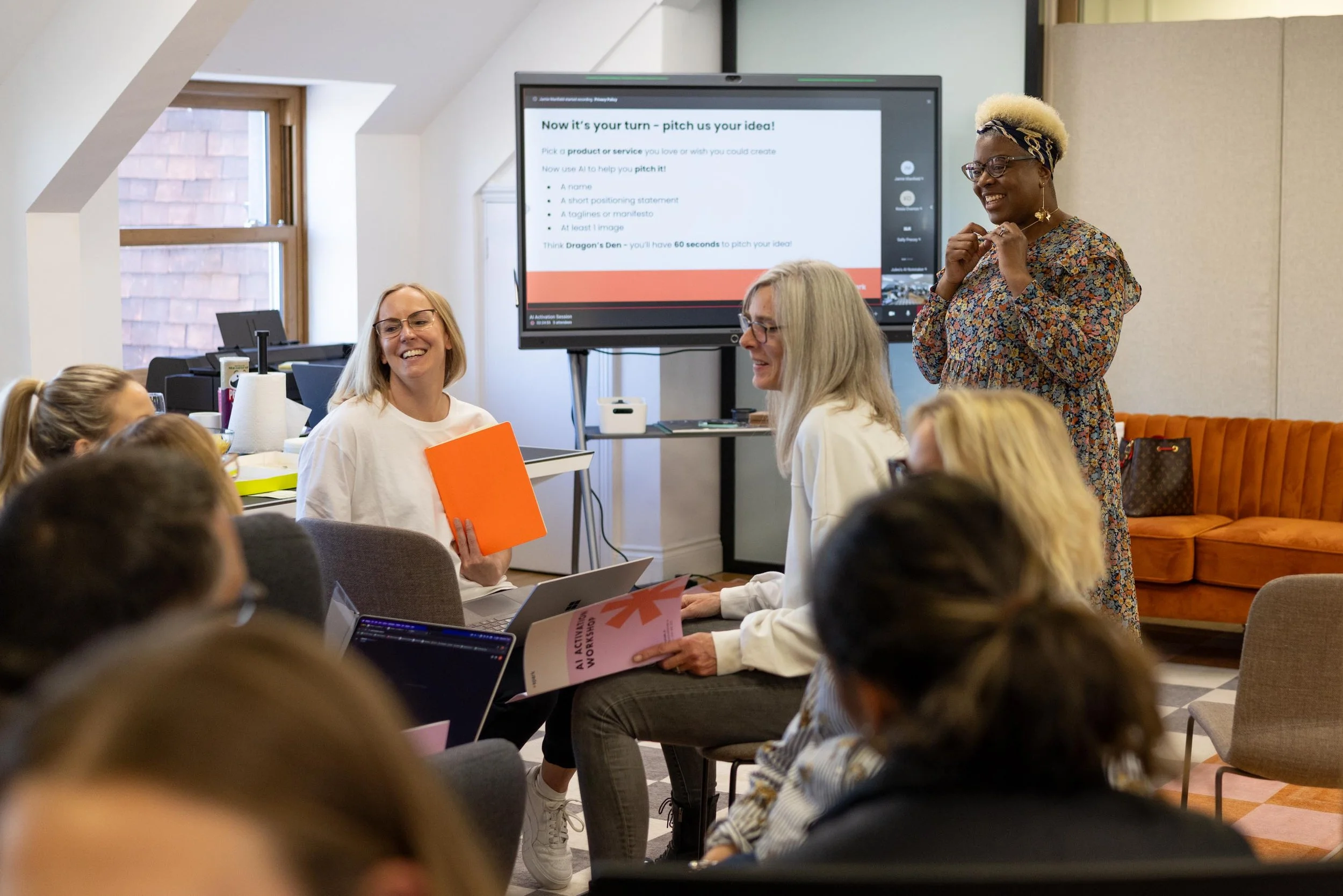 A diverse group of women attending a presentation in a conference room, with one woman standing and presenting, while others are seated, holding notebooks and laptops. There is a large screen displaying a presentation slide titled 'Now it's your turn - pitch us your idea!'