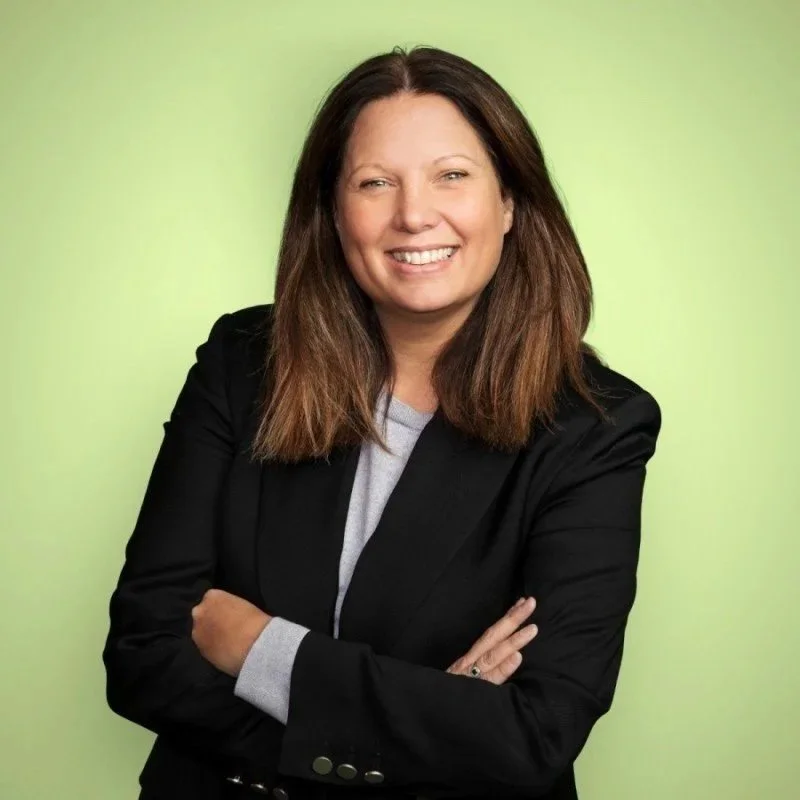 A woman with shoulder-length brown hair smiling and crossing her arms, wearing a black blazer and gray shirt, against a light green background.