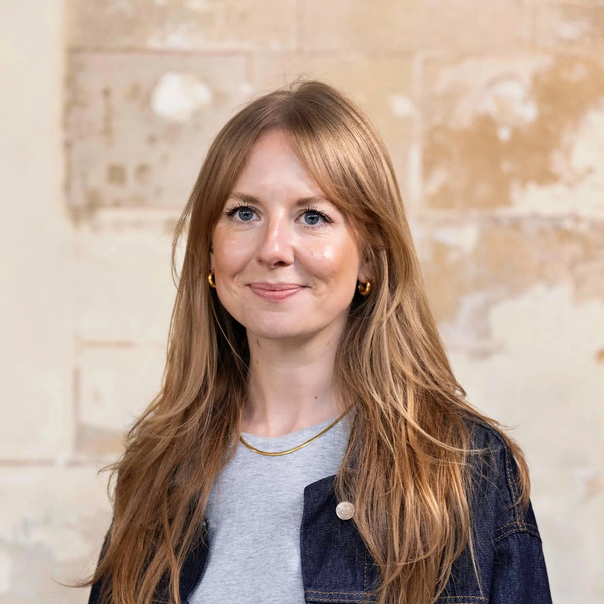 Close-up of a woman with wavy brown hair and a striped shirt smiling against a plain white background.