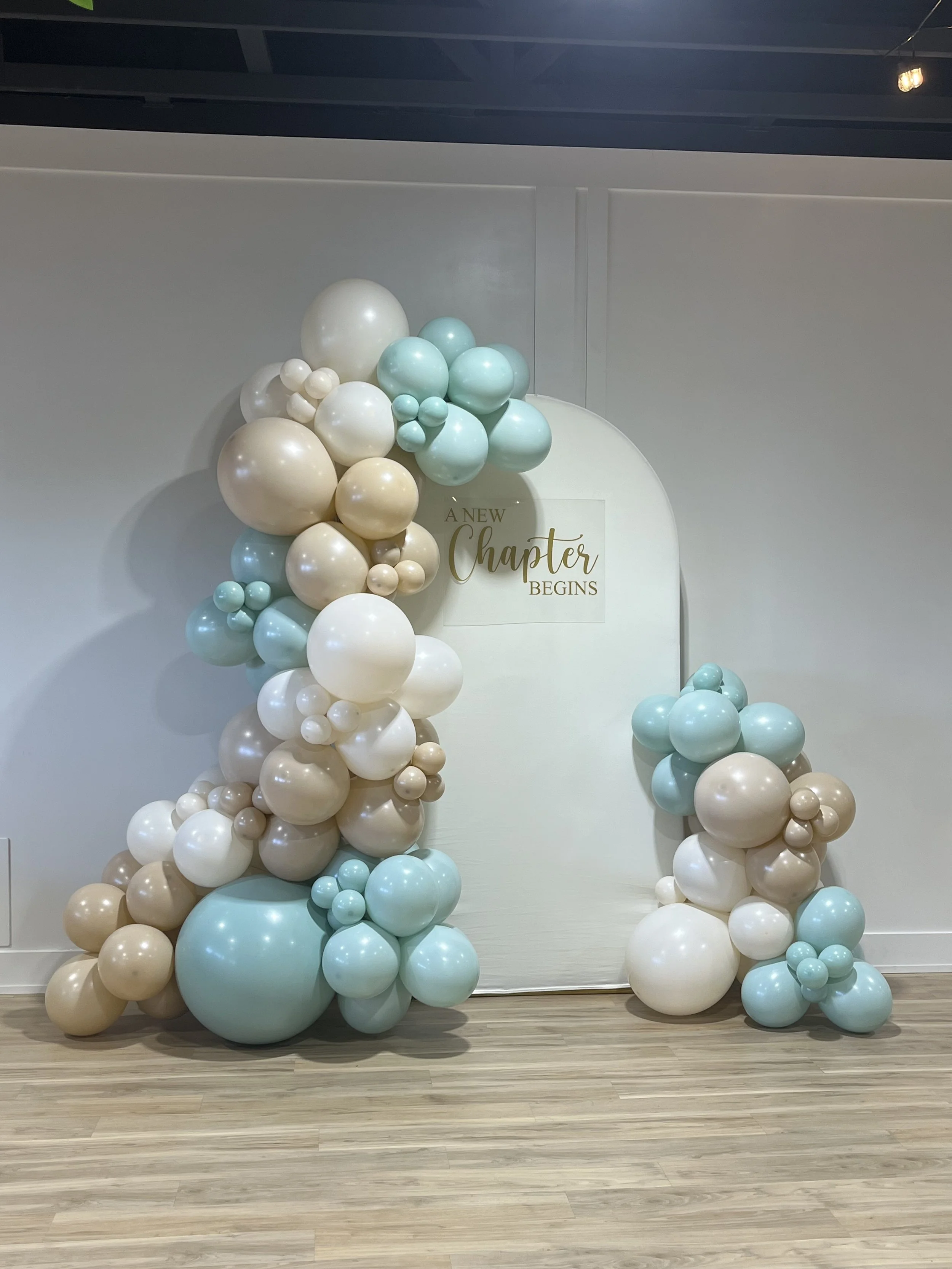 Balloon arch with pink, cream, and purple balloons decorated with pink cherry blossom branches, set against a plain wall with a round beige rug on the wooden floor.