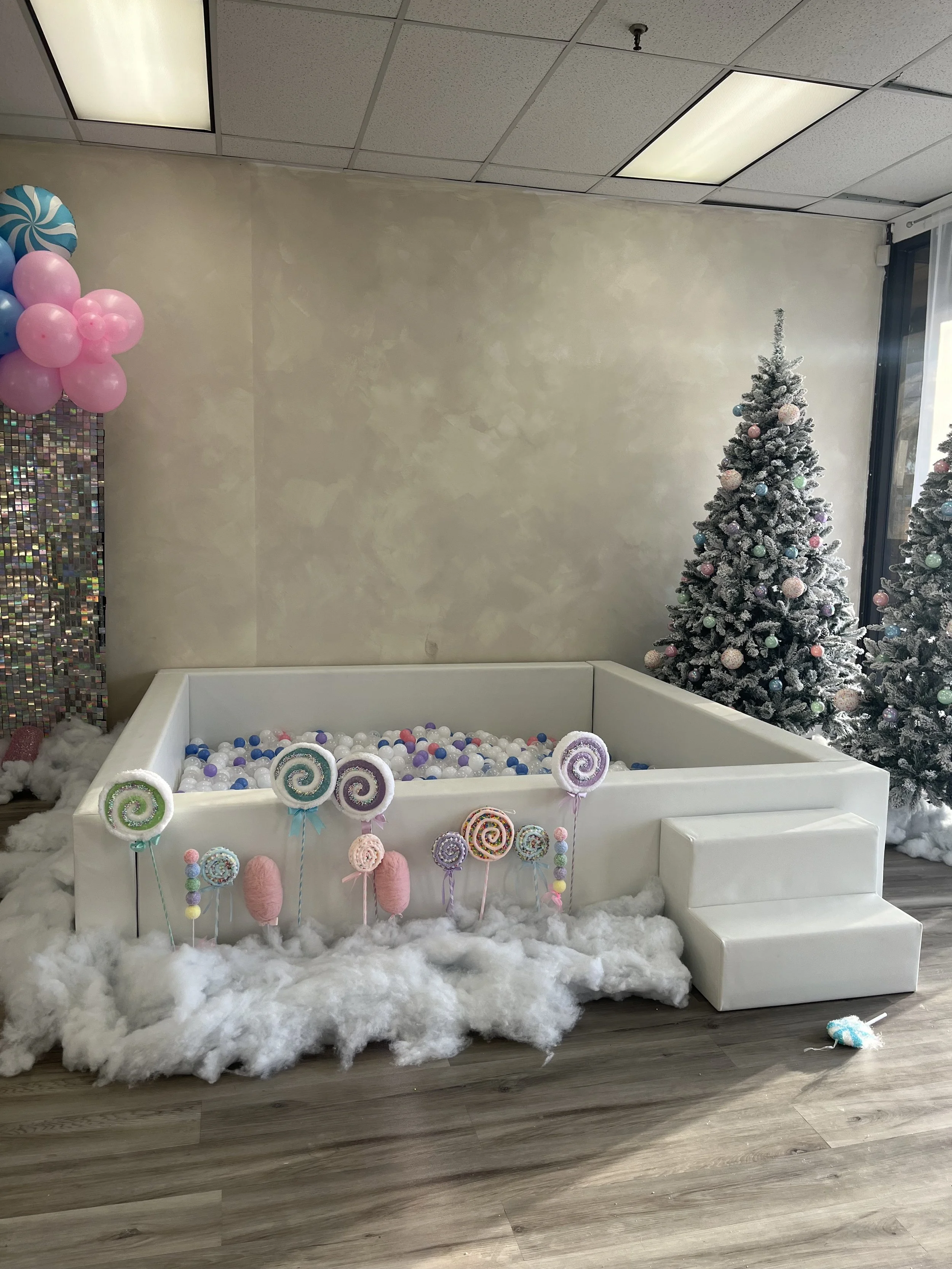 Decorated outdoor space with pink and white ball pit for a first birthday celebration, featuring a pink inflatable couch, a slide, and blocks spelling 'ONE' with floral and strawberry designs.