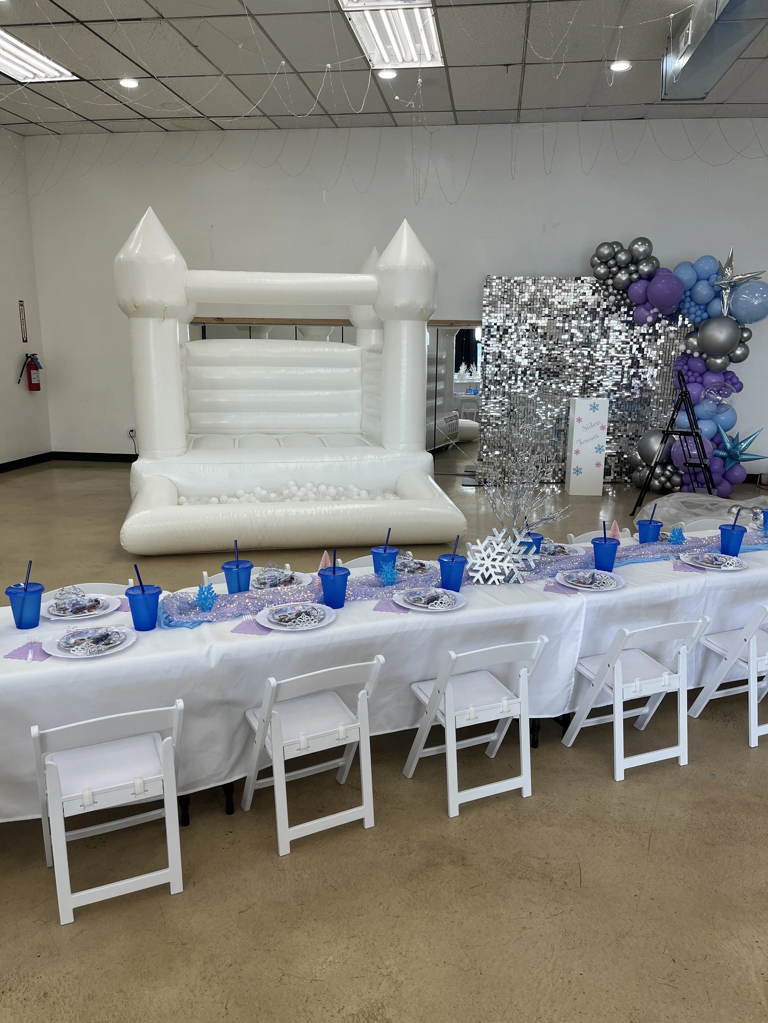 Two children playing on a large, white inflatable slide with a castle-like structure, decorated with pink and cream balloons, outdoors in a backyard with trees and lawn, while an adult supervises.