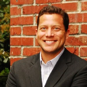 Headshot of a man with short hair smiling and wearing a black blazer with a white collared shirt