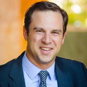 A headshot of a white middle aged man with short dark hair smiling in a suit and tie