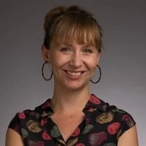 Headshot of a white woman with brown hair in a bun and bangs, smiling and wearing hoop earrings and a black blouse patterned with colorful plants