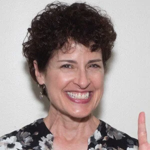 Headshot of a white woman with short, dark, curly hair smiling and wearing a dark floral blouse